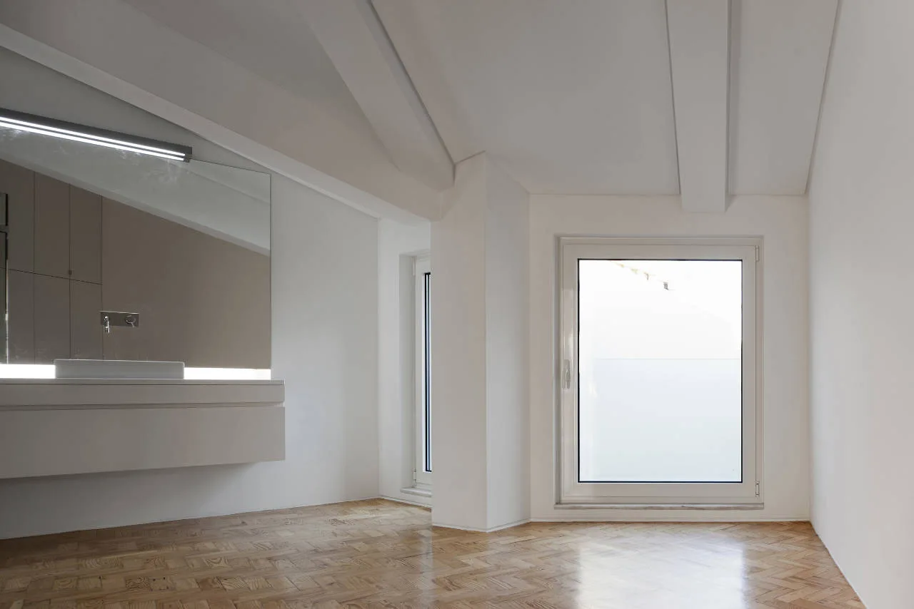 A minimalist white bathroom featuring a floating vanity, a backlit rectangular mirror, and light wood herringbone flooring under structural ceiling beams.