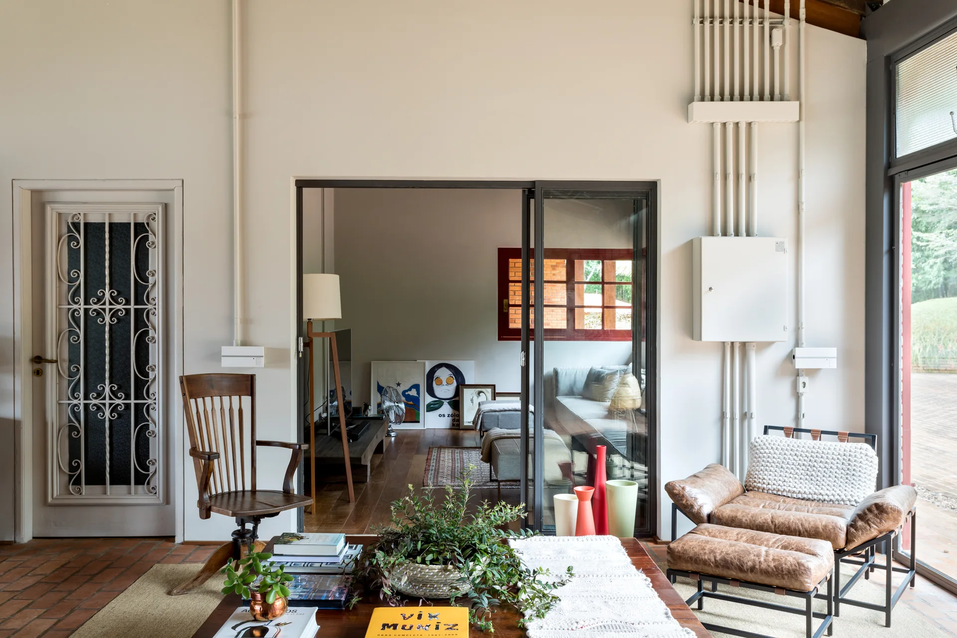 A sunlit living room features a leather lounge chair, a wooden coffee table, and black-framed glass sliding doors opening to an adjacent space.