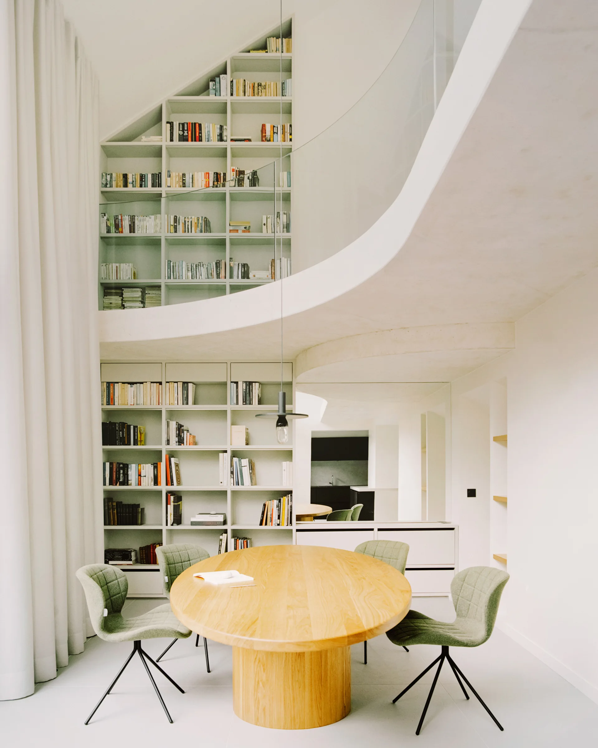 A minimalist dining area featuring an oval wooden table and green chairs under a curved white mezzanine and tall built-in bookshelves.