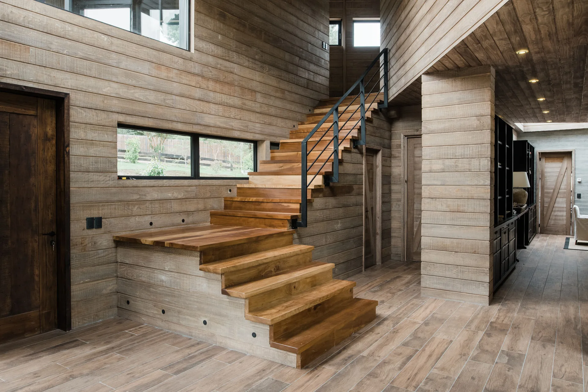 A rustic home entryway features extensive horizontal wood wall paneling, a wooden staircase with a black metal railing, and matching wood floors.