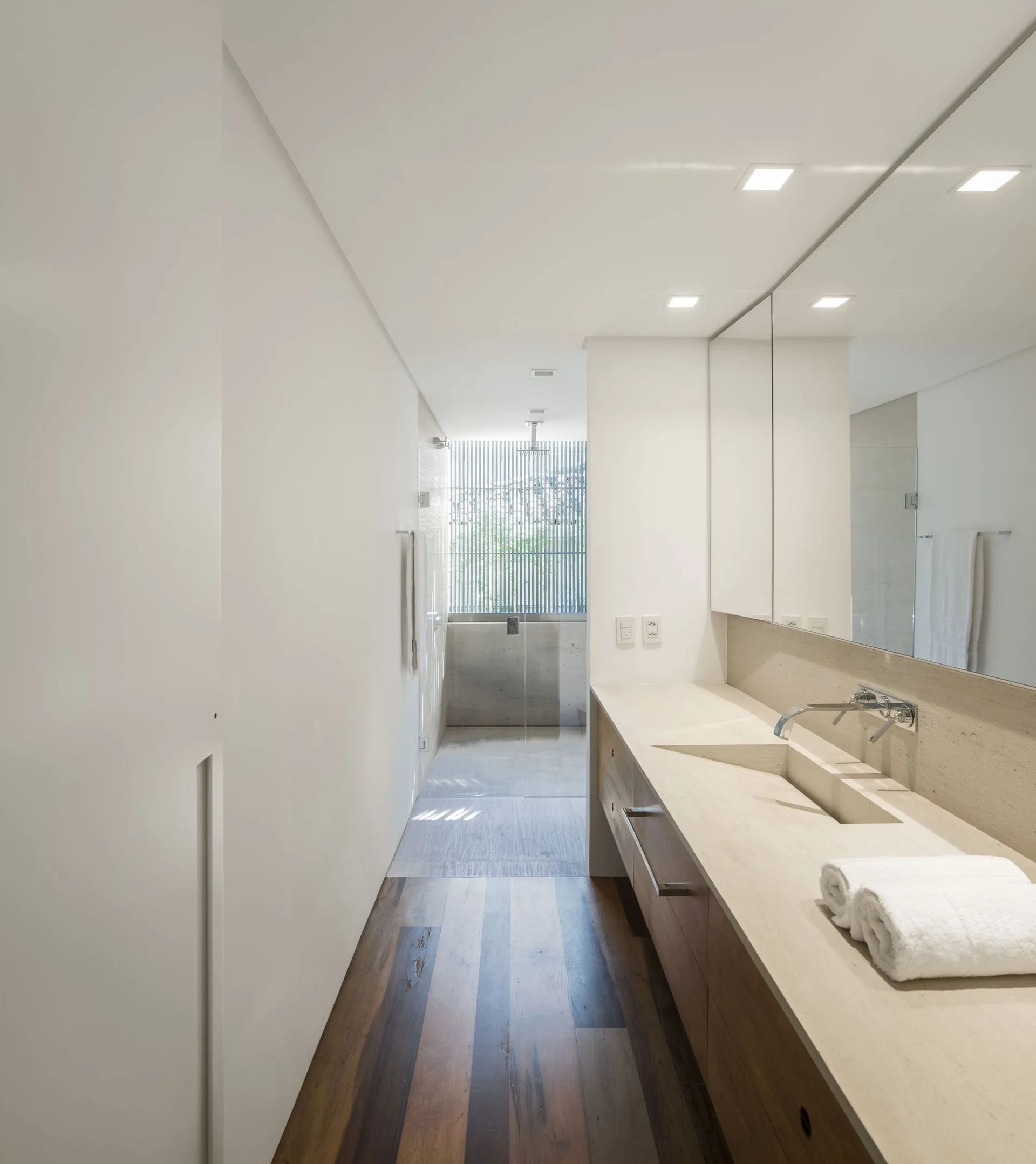 A minimalist narrow bathroom featuring a long stone vanity with an integrated sink, dark wood floors, and a glass shower in the background.
