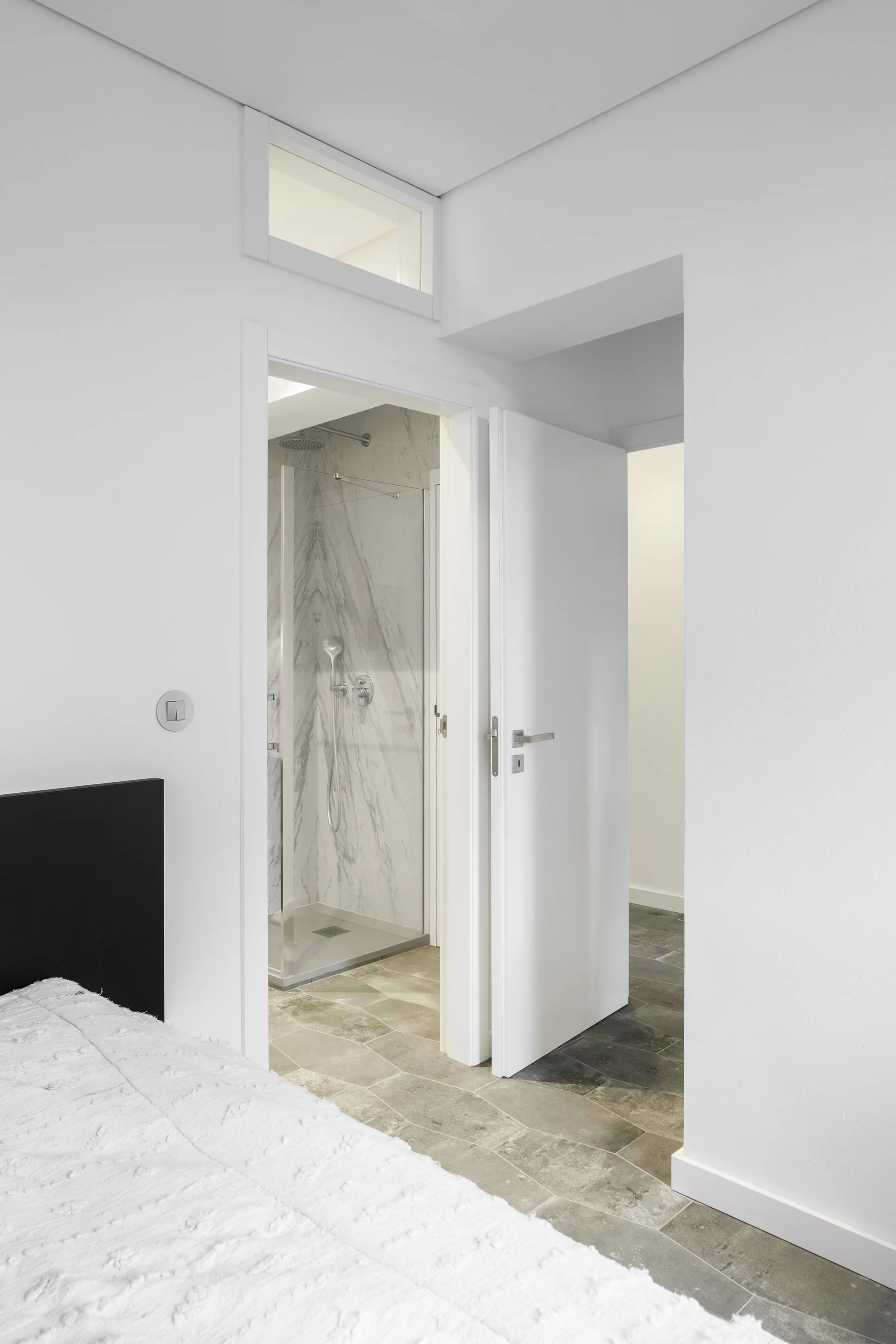 A view from a bedroom with a white bedspread into an adjacent bathroom featuring a marble-walled shower and a transom window above the door.