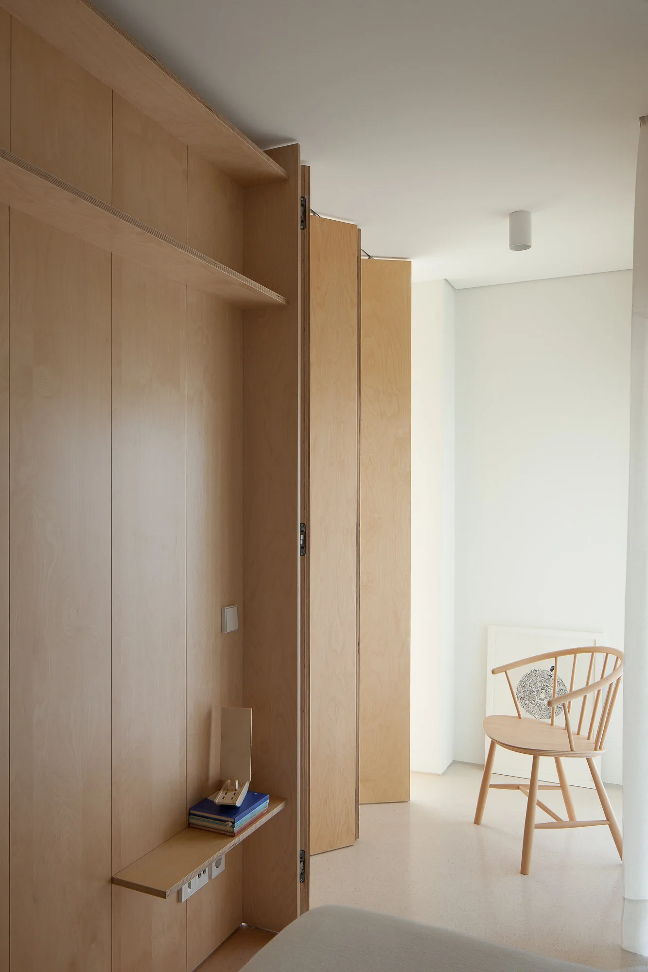 A minimalist room featuring floor-to-ceiling light wood cabinetry with built-in shelves and a light wood spindle chair against a white wall.