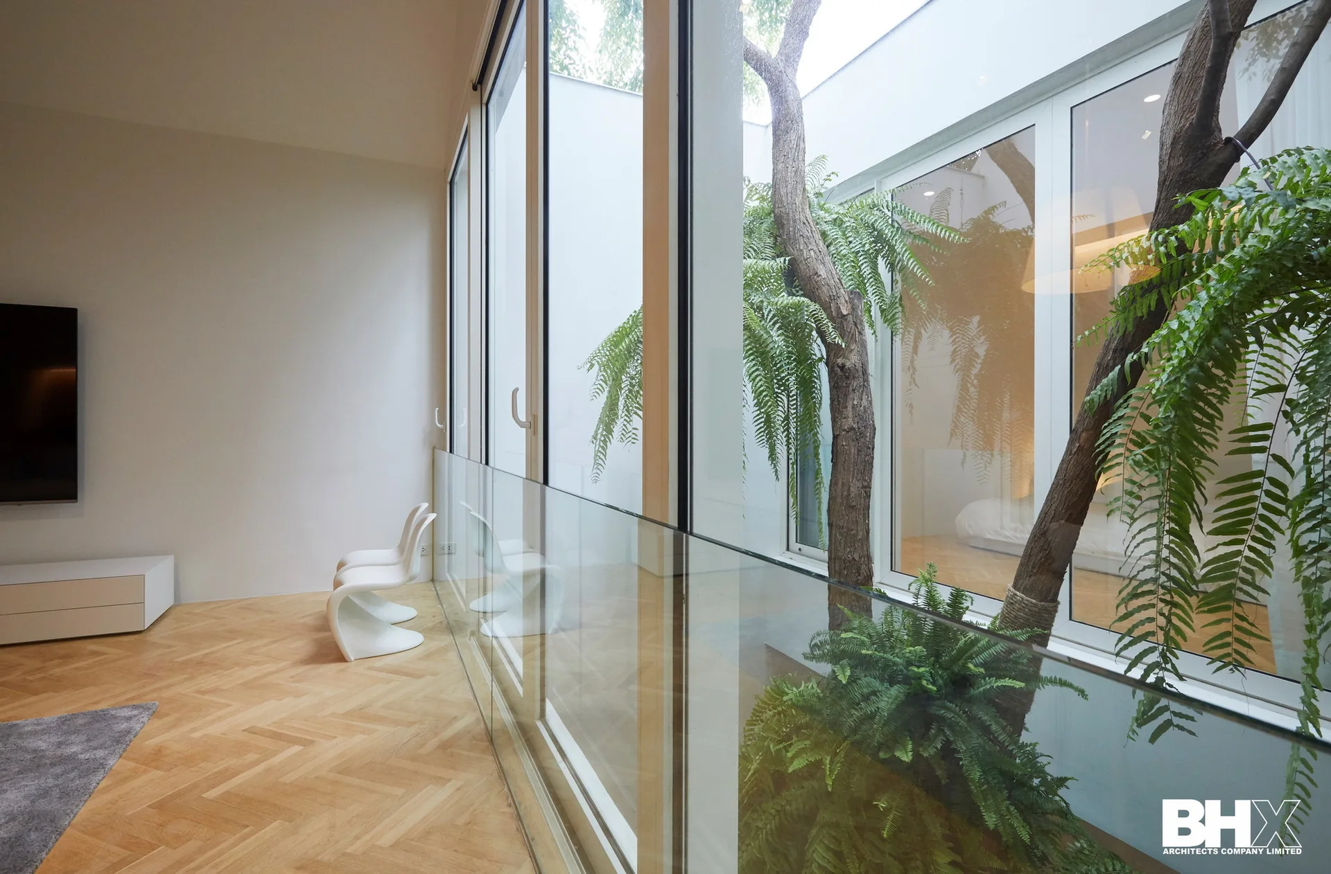 A minimalist living room with herringbone wood floors features large glass walls overlooking an internal courtyard with lush green trees.