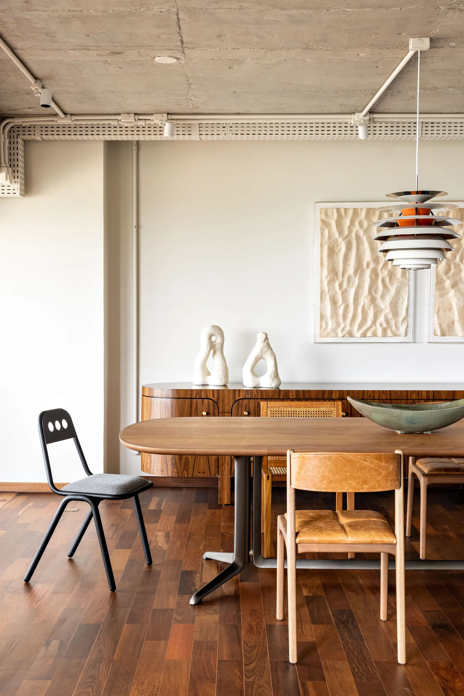 A modern dining room features a wooden dining table, a curved wood sideboard, and mismatched chairs beneath an exposed concrete ceiling.