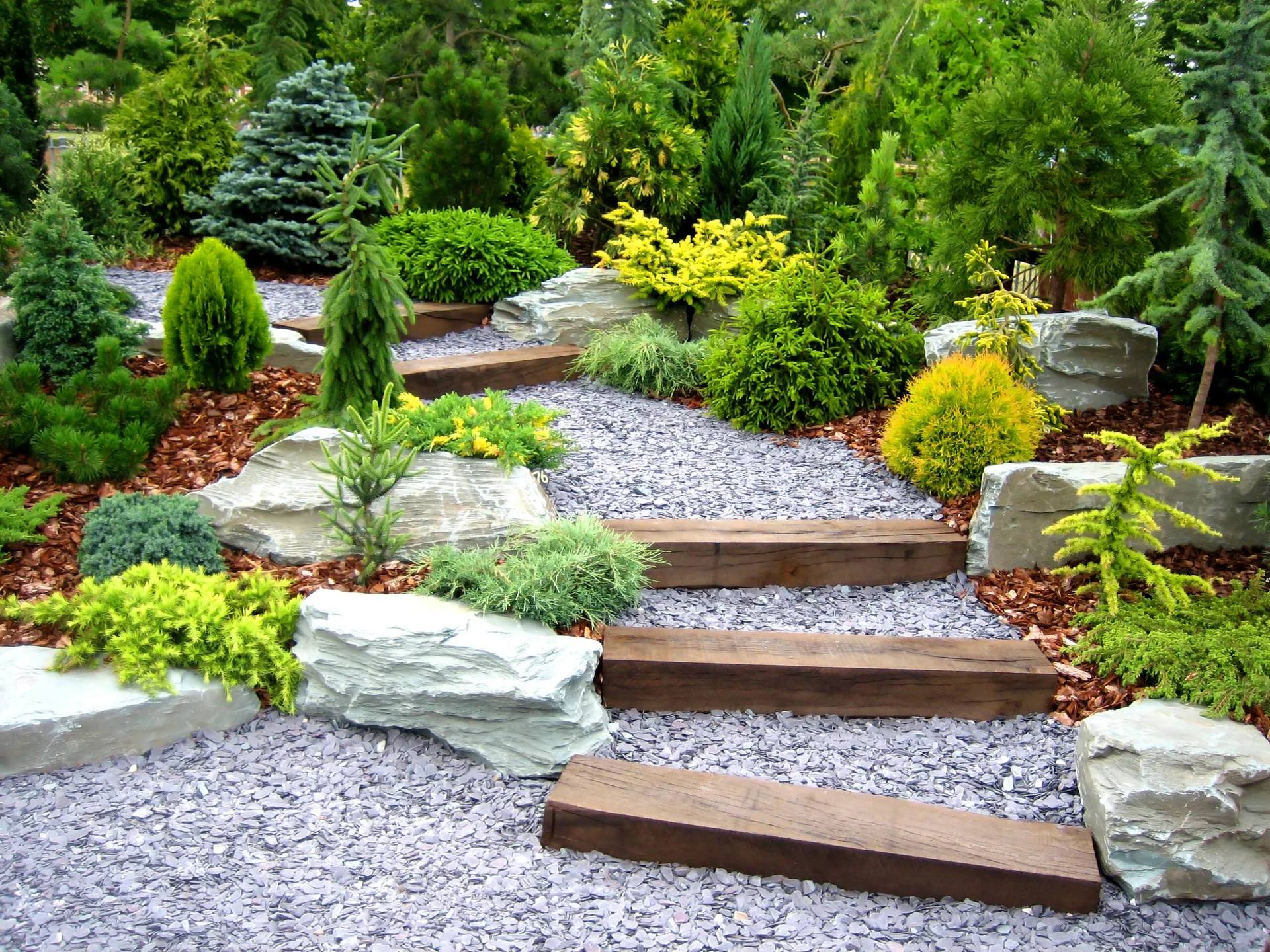 A sloped garden landscape featuring a grey gravel path with dark wood steps, surrounded by large grey boulders and green and yellow conifers.