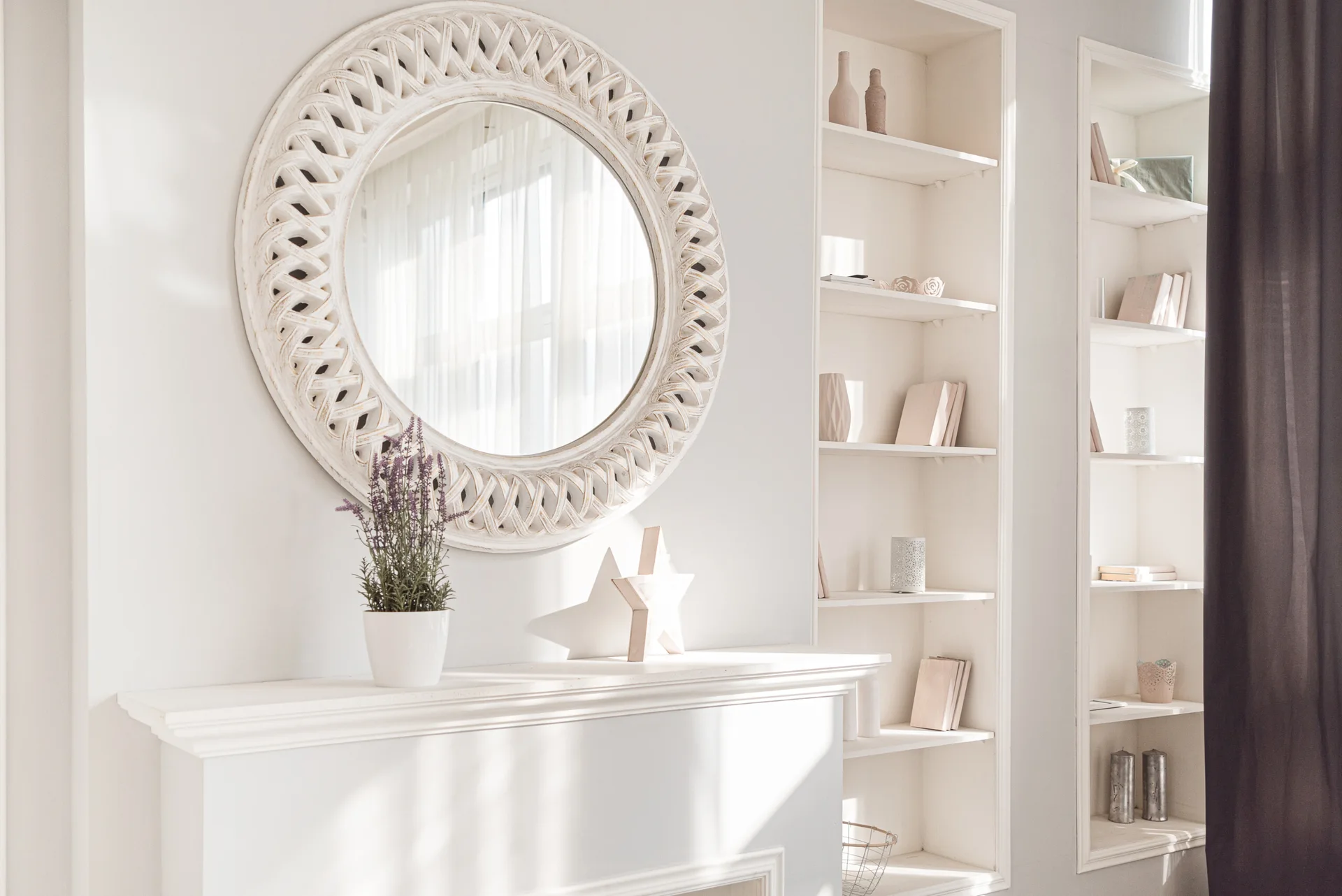 A white fireplace mantel features a round decorative mirror and is flanked by tall recessed shelves styled with neutral books and pottery.