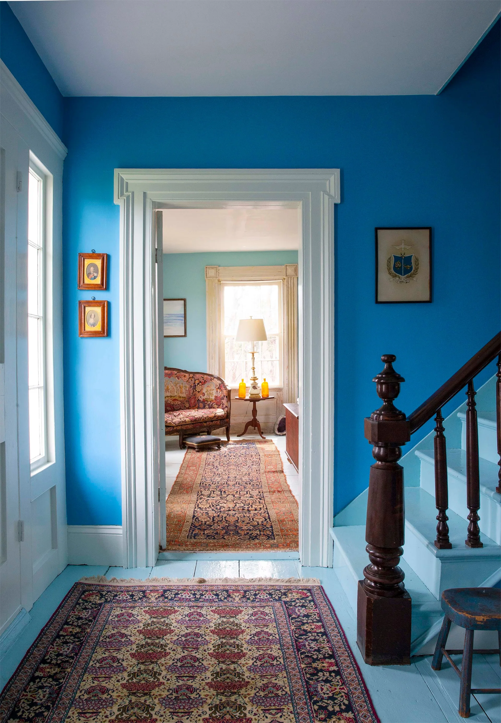 A bright blue residential hallway features painted wood floors, a classic staircase with a dark wood handrail, and a wide doorway framing a sitting room.