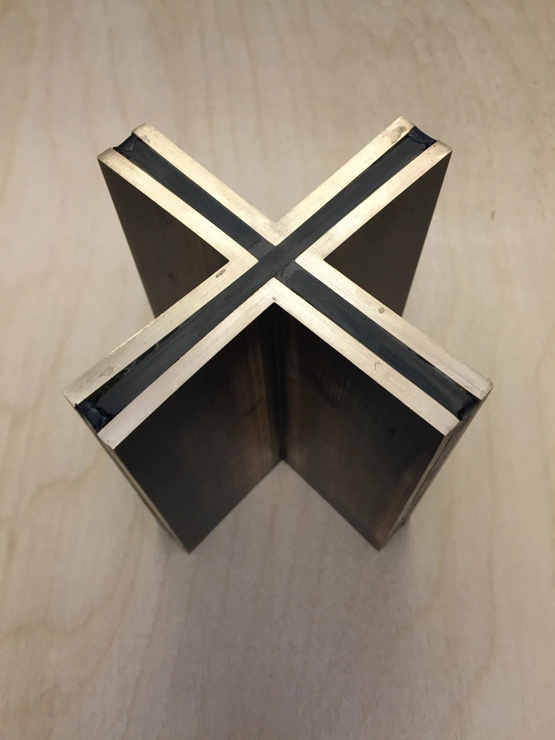 A cross-shaped architectural joint sample of layered brass and black composite stands upright on a light plywood table.