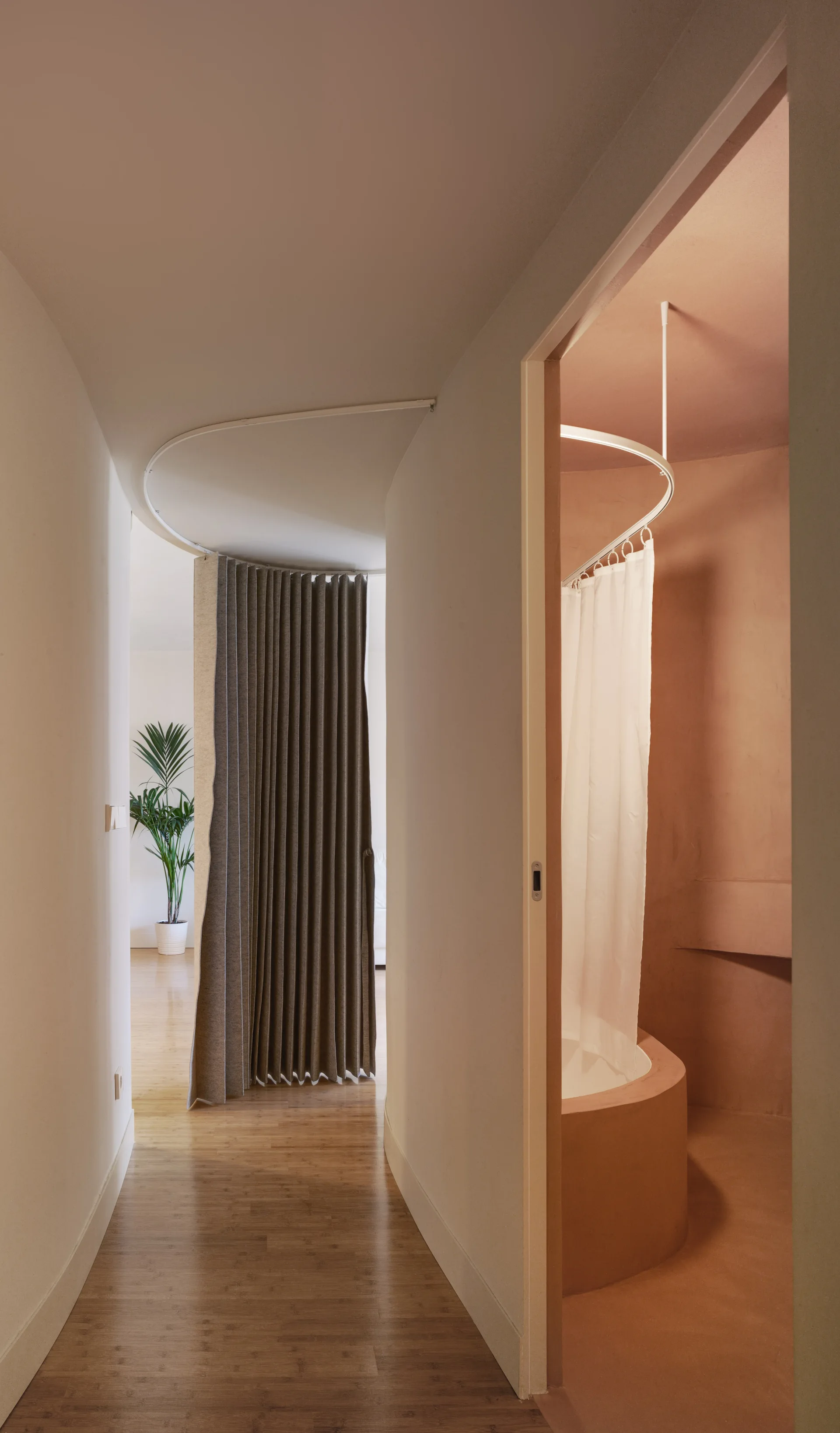 A curved white hallway features a gray pleated curtain room divider leading past a terracotta bathroom with a circular tub.