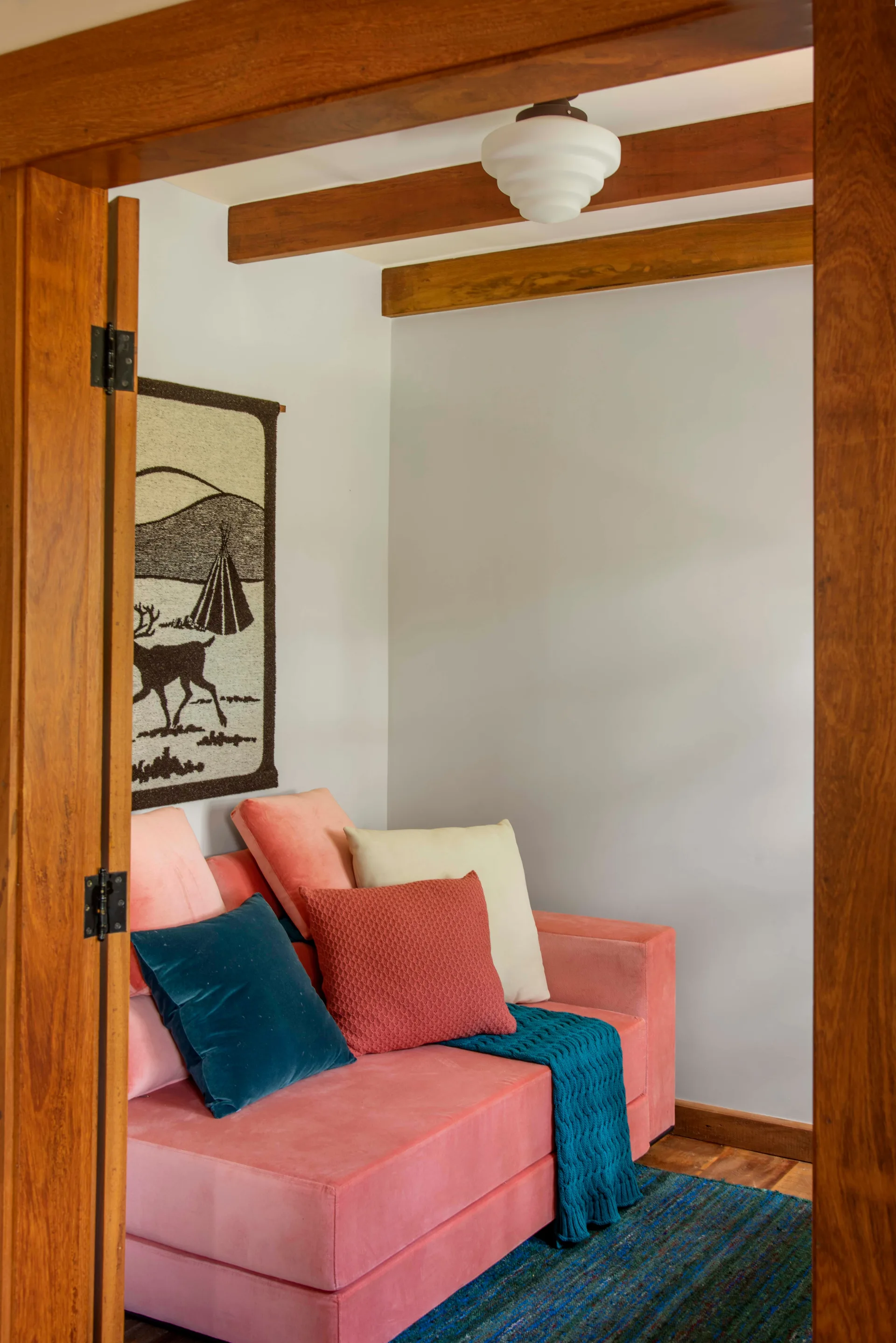 A view through a wooden doorway into a room featuring a pink velvet sofa with colorful pillows, exposed ceiling beams, and a woven wall hanging.