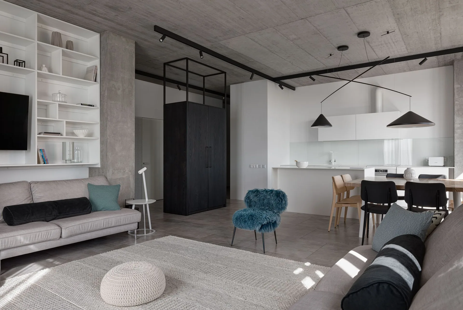 An open-plan living room and kitchen featuring two gray sofas, an exposed concrete ceiling, built-in white shelving, and a fuzzy blue accent chair.