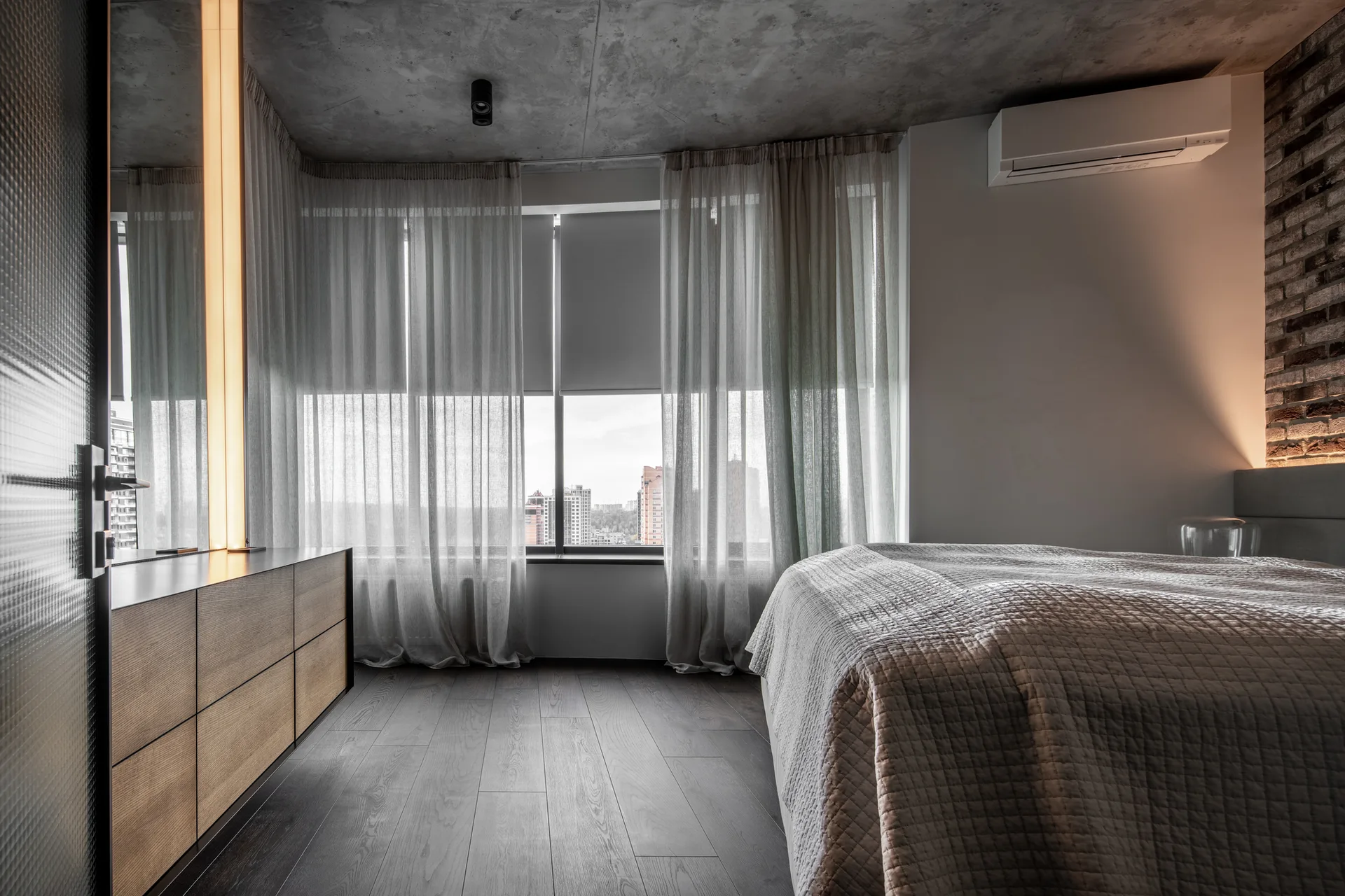 An industrial bedroom with an exposed concrete ceiling, a brick accent wall, a quilted bed, and a wooden dresser near sheer curtains.