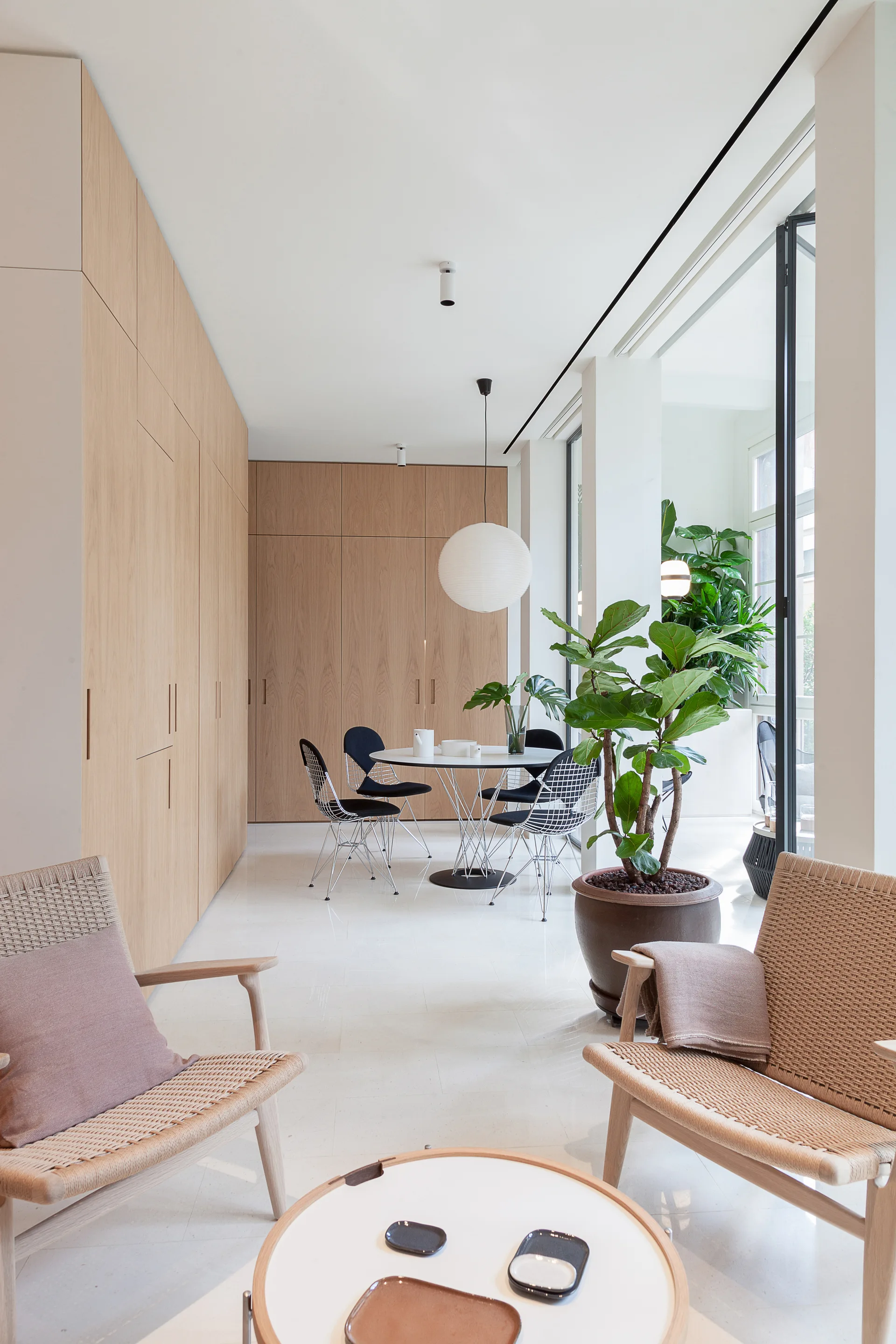 A bright minimalist interior showing woven armchairs in the foreground and a dining table with wire chairs next to seamless wood wall cabinets.