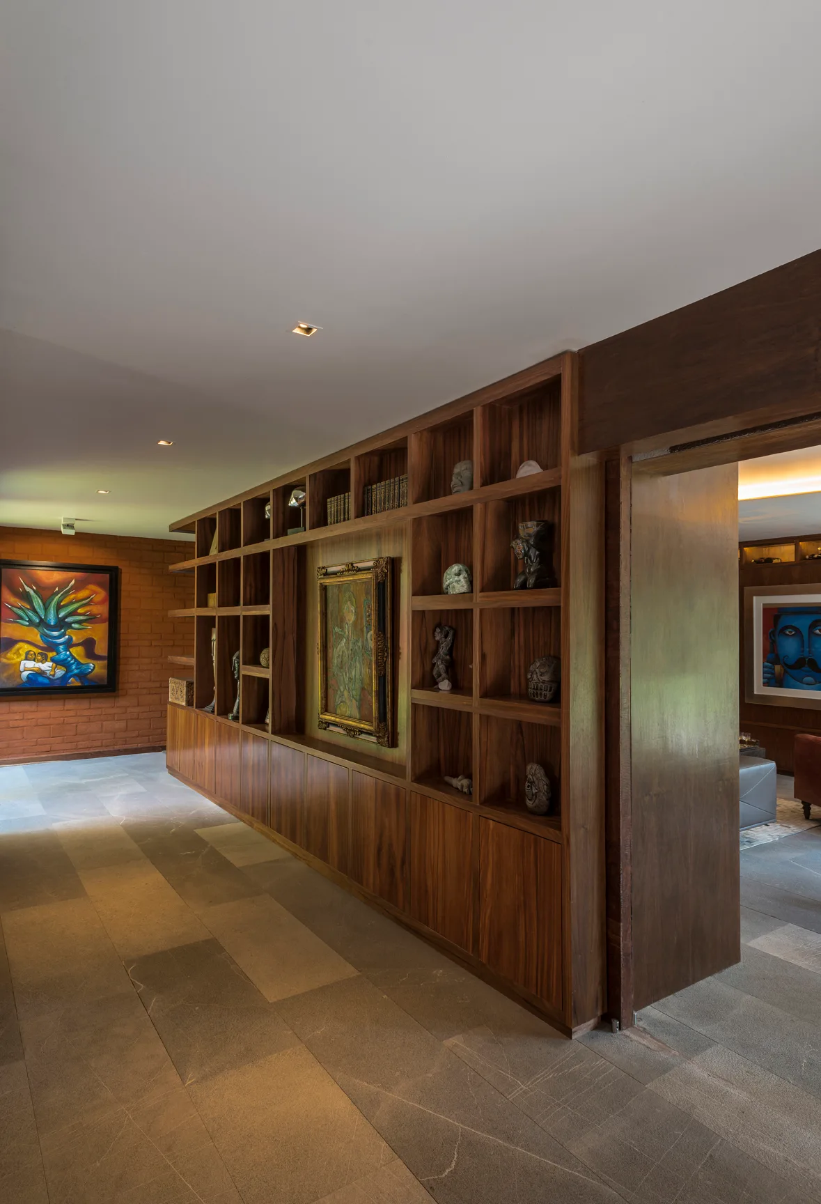 A wide hallway featuring a custom wooden shelving unit partition, stone tile flooring, and a red brick wall in the background.