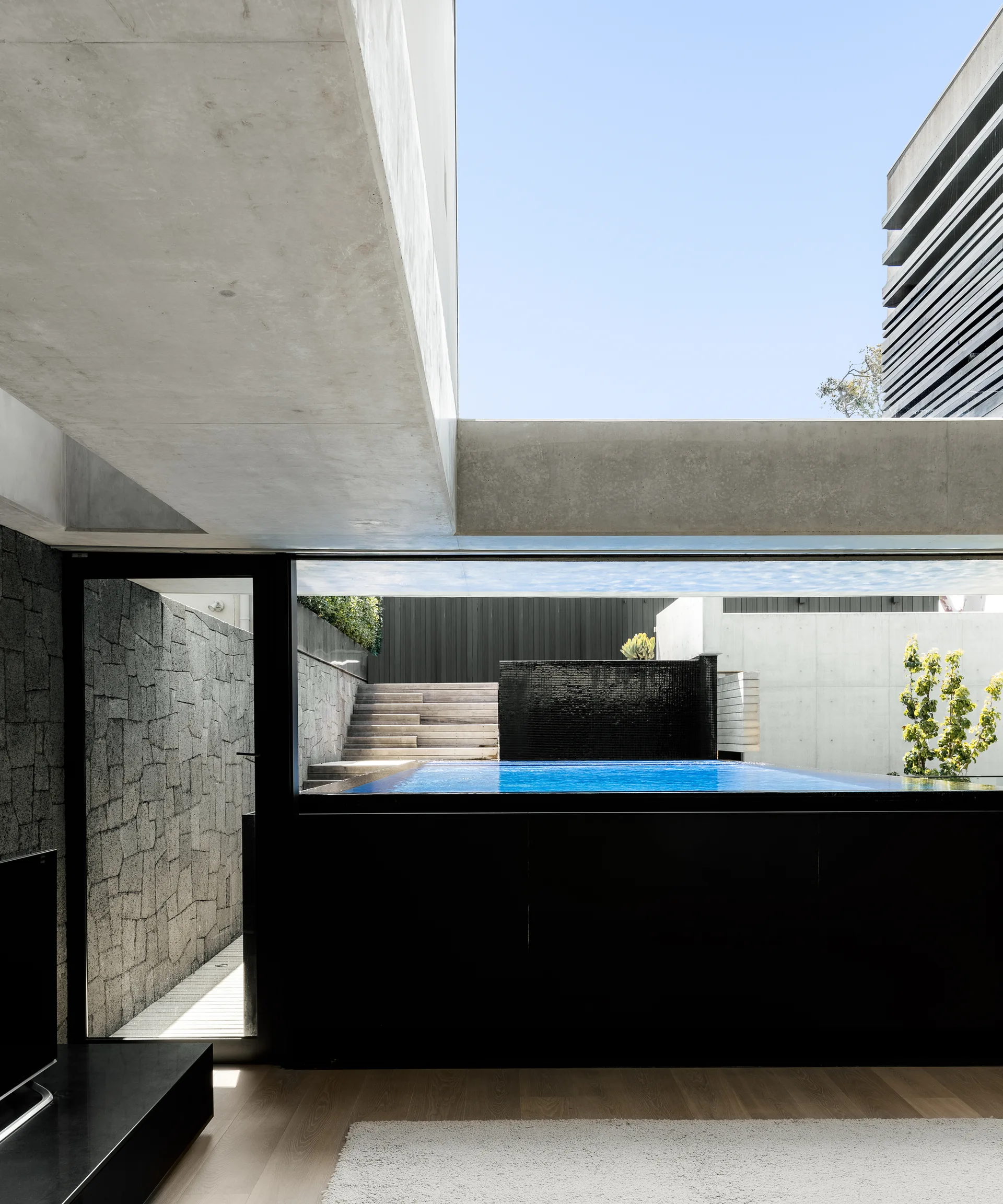 An interior room with wood floors looks out through a large window toward an elevated blue swimming pool, concrete beams, and a stone wall.