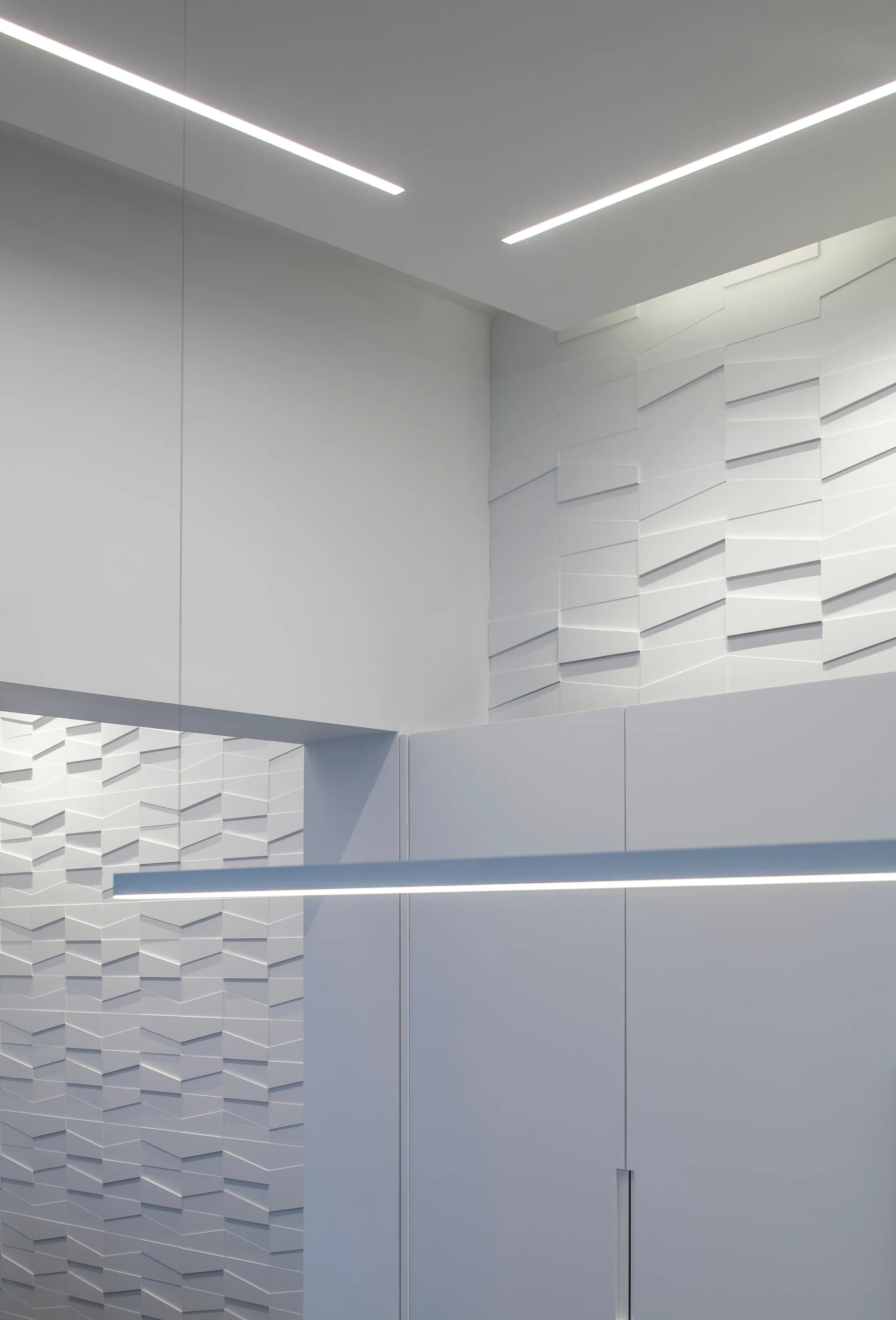 A modern minimalist white interior features geometric three-dimensional wall paneling, a suspended linear pendant light, and recessed ceiling lights.
