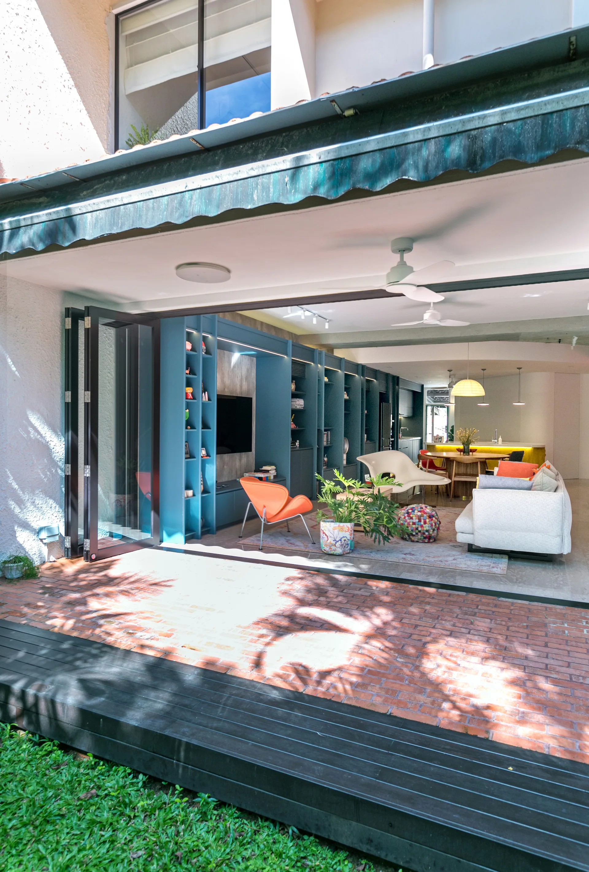 A modern living room features a blue built-in shelving unit and an orange accent chair, opening onto a red brick patio through folding glass doors.