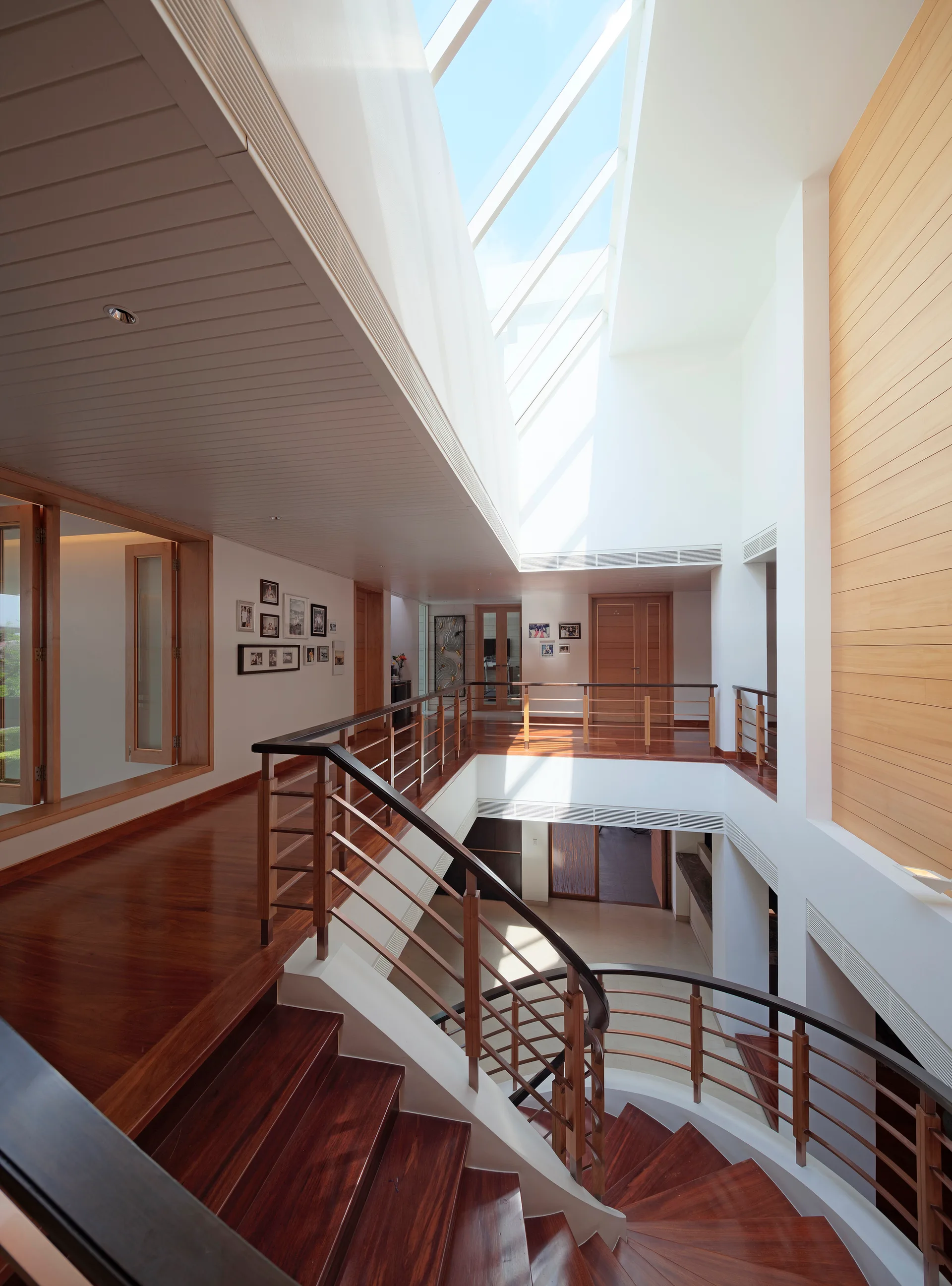 A multi-level hallway features a curved wooden staircase with dark steps, metal railings, polished wood floors, and a large skylight overhead.
