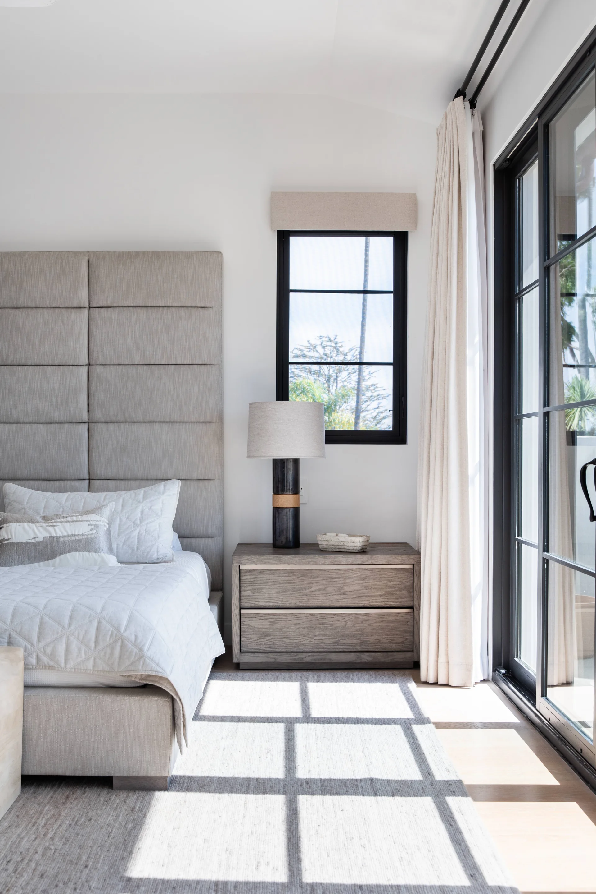 A contemporary bedroom featuring a tall upholstered headboard, a wooden bedside drawer, and a large black-framed sliding glass door.