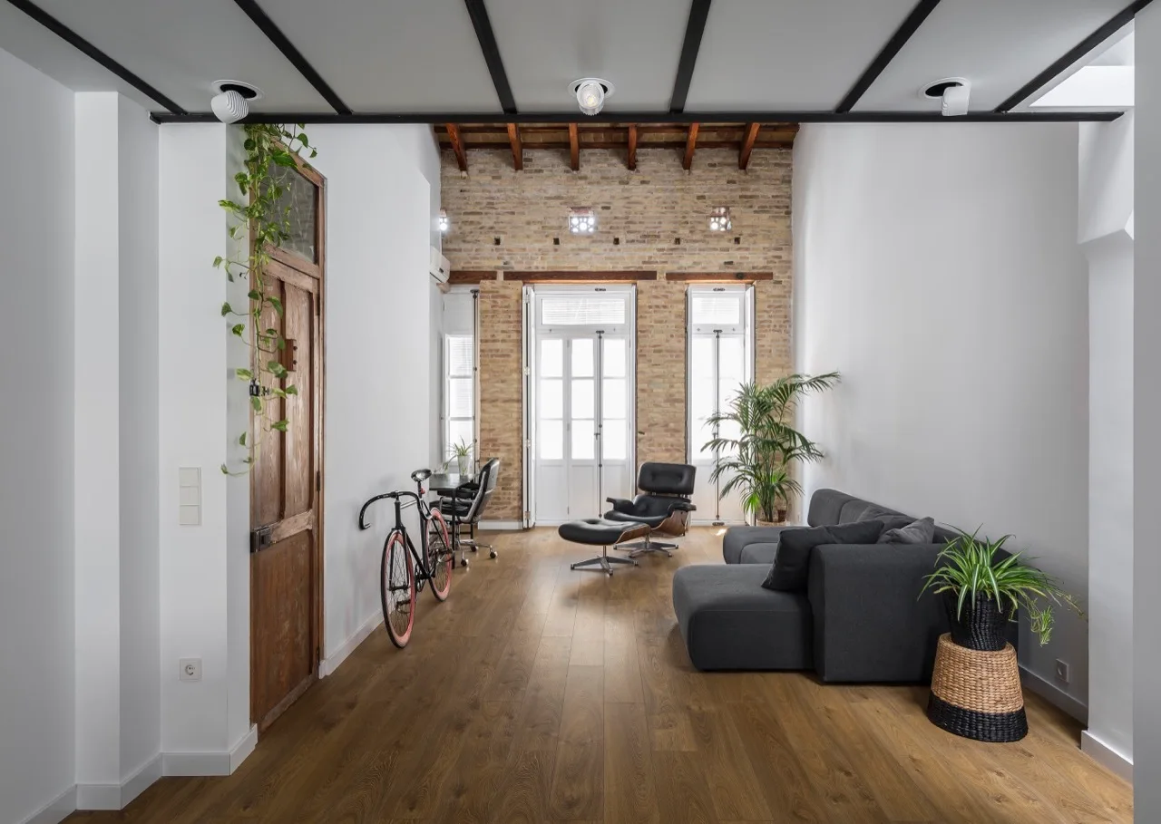 A spacious loft living room featuring a charcoal sectional sofa, a black leather lounge chair, hardwood flooring, and a textured exposed brick wall.