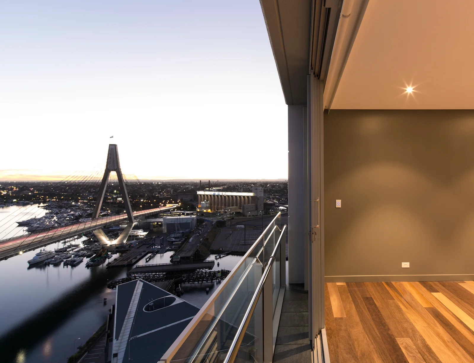 An elevated apartment balcony with a glass and metal railing overlooks a harbor and suspension bridge at twilight next to an indoor wood floor.