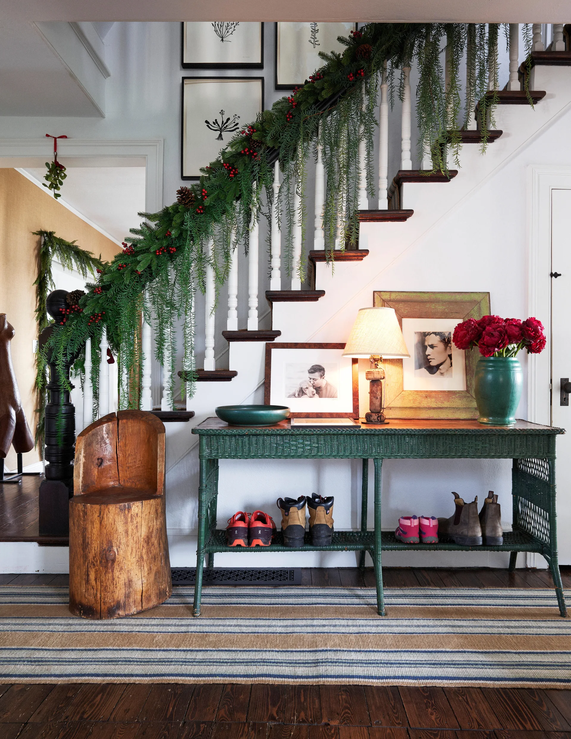 A green wicker console table sits beneath a staircase draped in a lush berry garland, flanked by a carved wood stump chair.