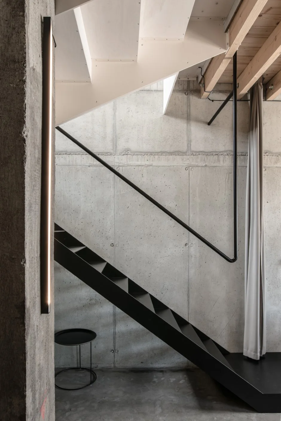 A black metal staircase stands against a raw exposed concrete wall, paired with a vertical linear light sconce and a grey fabric curtain.
