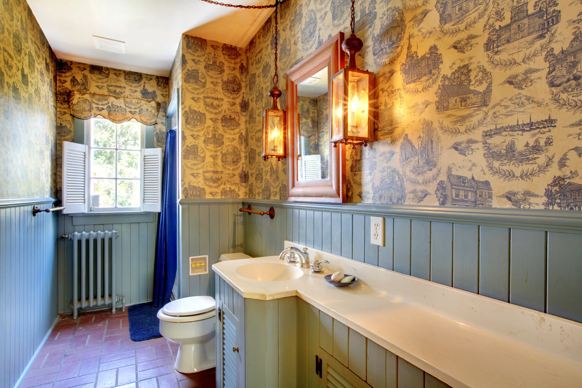 A traditional bathroom featuring sage green wainscoting below scenic blue and cream toile wallpaper, with a wooden vanity and hanging lantern lights.