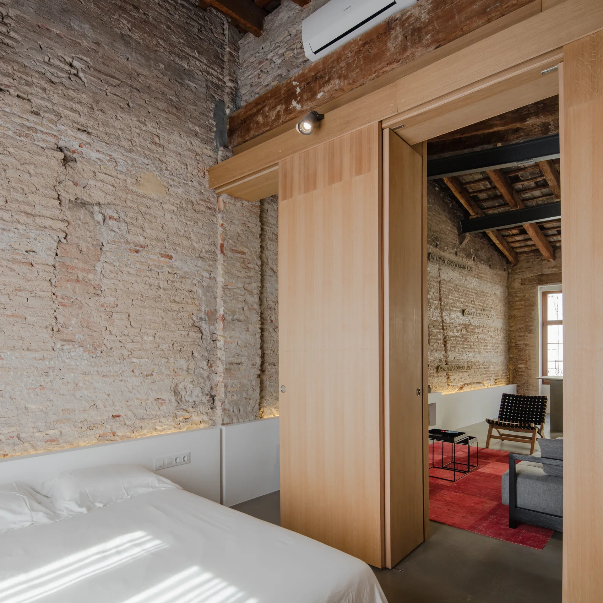 A bedroom with an exposed brick wall and white bed features a large wooden sliding partition door opening to a living area.