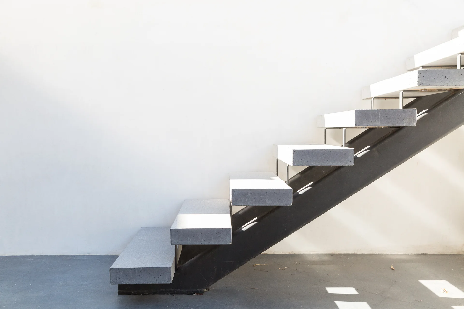 A minimalist staircase with thick grey concrete treads supported by a dark metal stringer against a clean white wall.