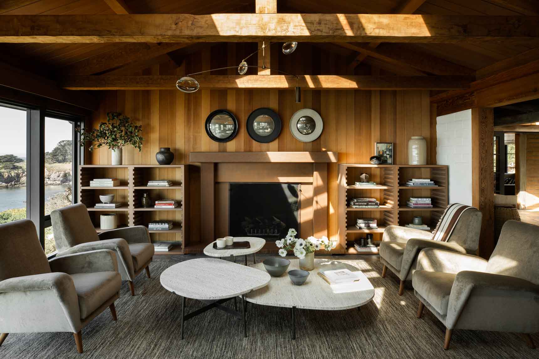 A wood-paneled living room features four grey armchairs arranged around nested stone coffee tables in front of a fireplace.