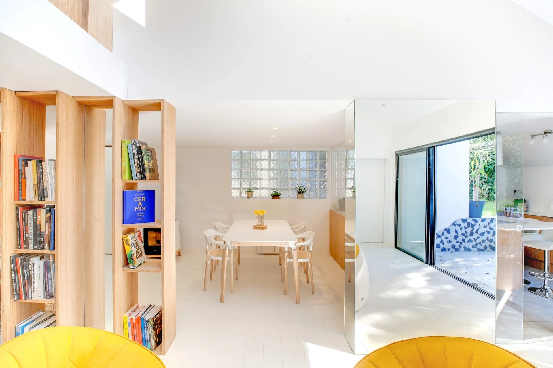 A minimalist white dining room features a light wood table, surrounding chairs, a tall wooden bookshelf partition, and a glass block window.