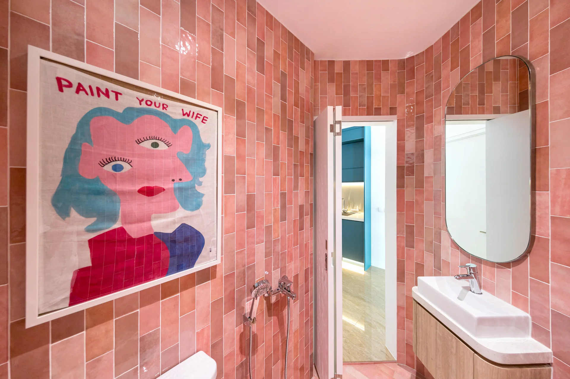 A small bathroom features pink vertical tile walls, a white sink on a wood vanity, an oval mirror, and a colorful framed art piece.