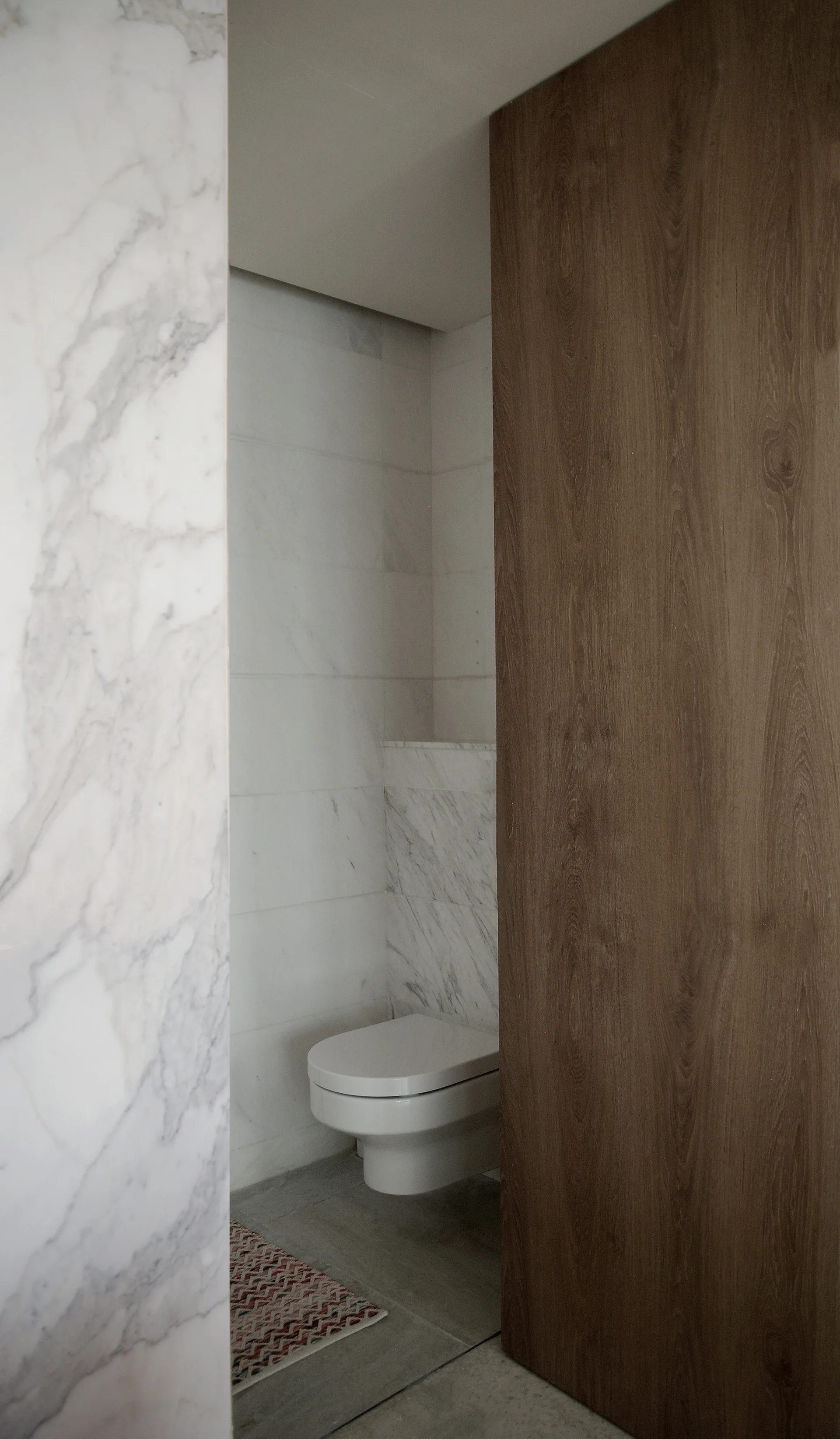A narrow view of a minimalist powder room with a white wall-mounted toilet, marble-tiled walls, and a large dark wood accent panel.