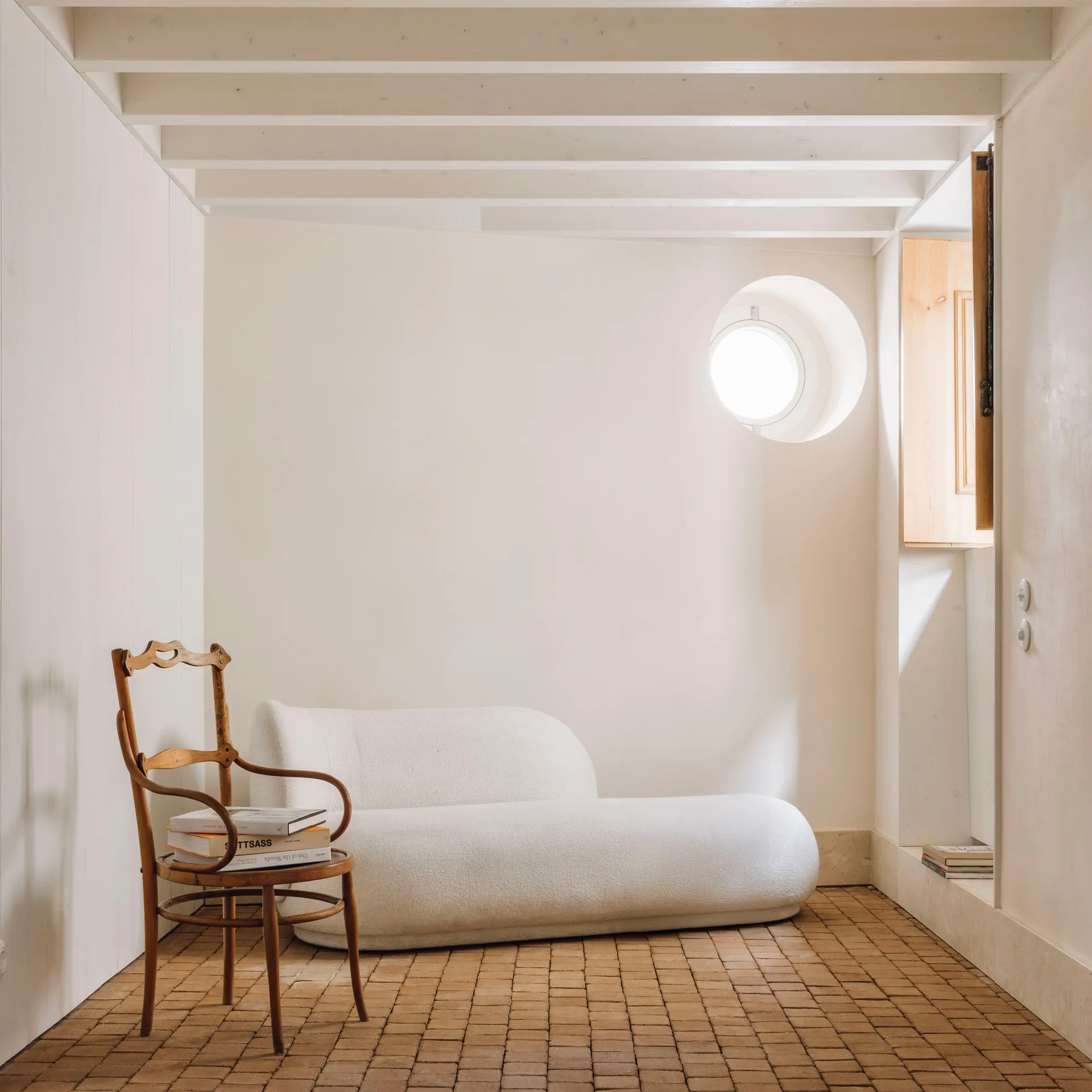 A minimalist room features a white curved sofa, a vintage wooden chair with books, a round window, and a terracotta brick floor.