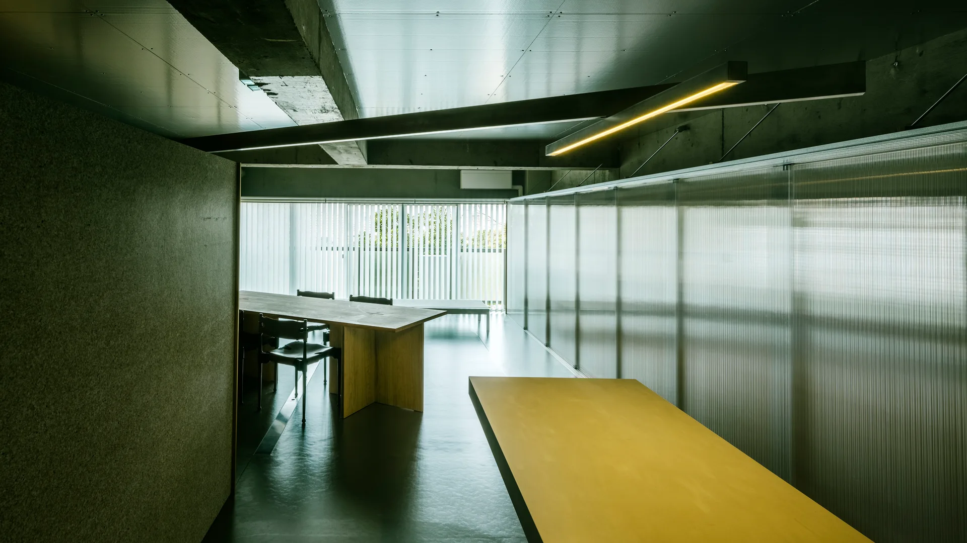A minimalist dining room features a long wooden table, black chairs, a translucent corrugated plastic partition wall, and an exposed concrete ceiling.