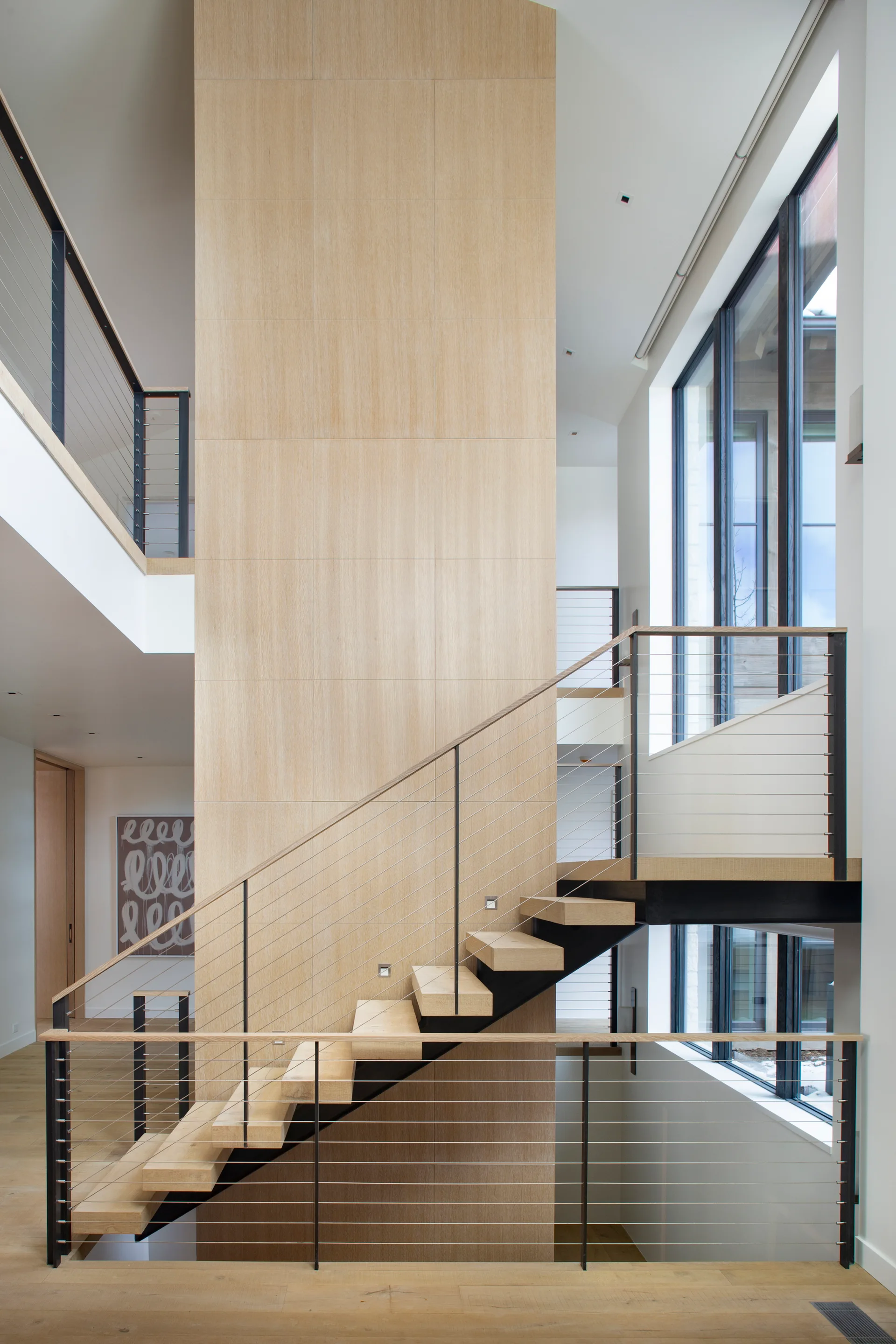 A modern multi-level staircase features light wood floating treads, thin black metal cable railings, and a tall wood-paneled central column.