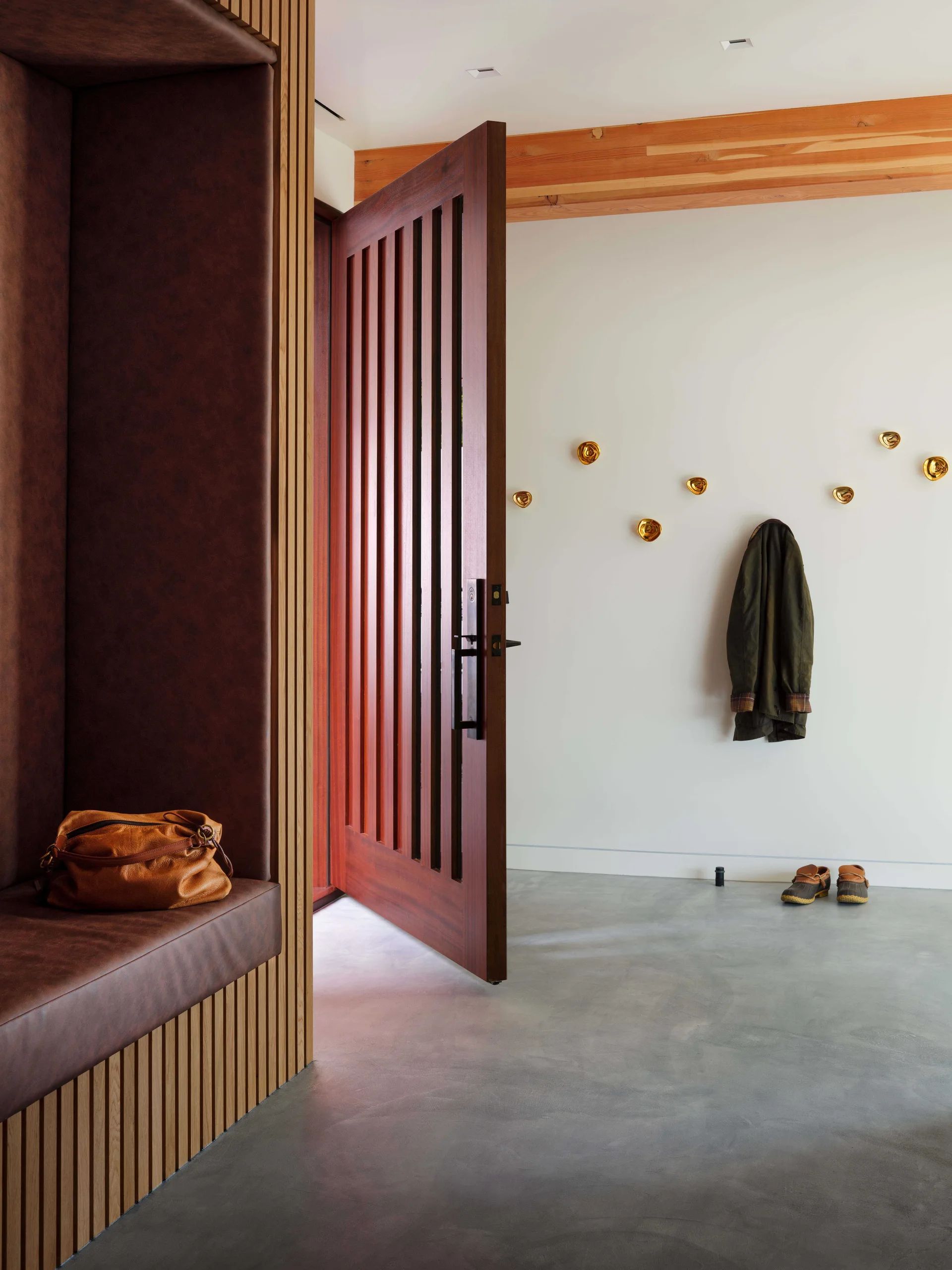A modern entryway features an open wood-slatted door, a built-in brown leather alcove bench, and decorative gold hooks on a white wall.