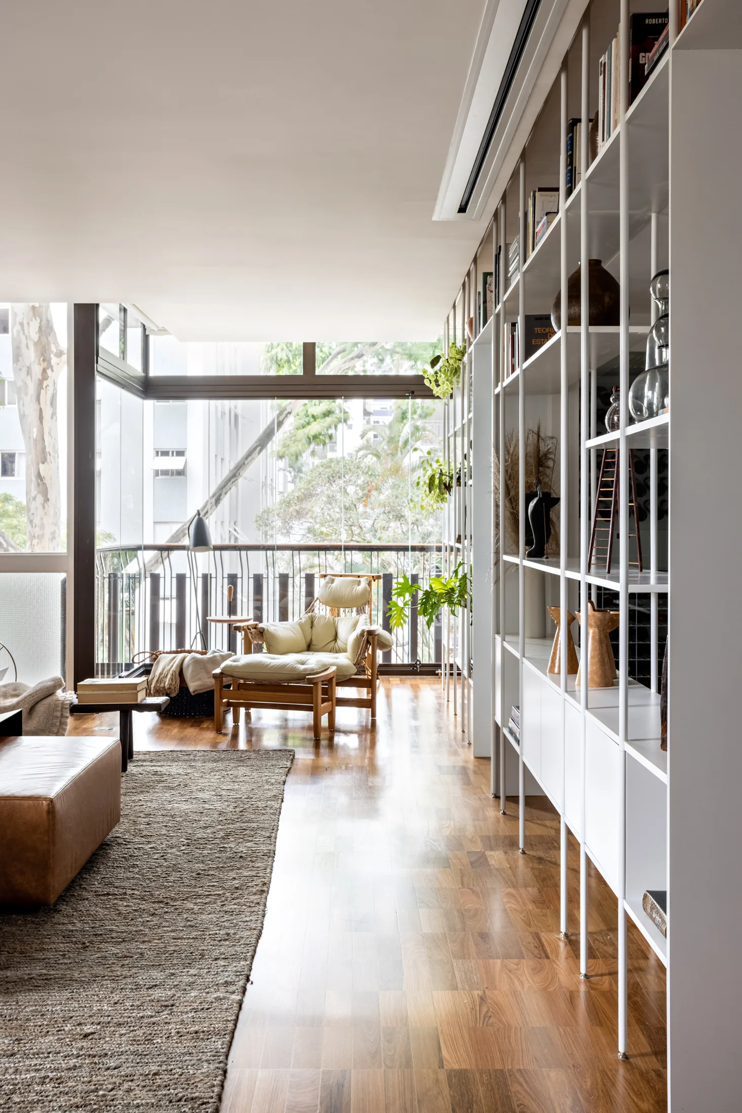 A bright living room features a tall white open bookshelf, a cream armchair with an ottoman, and a glass wall looking out to greenery.