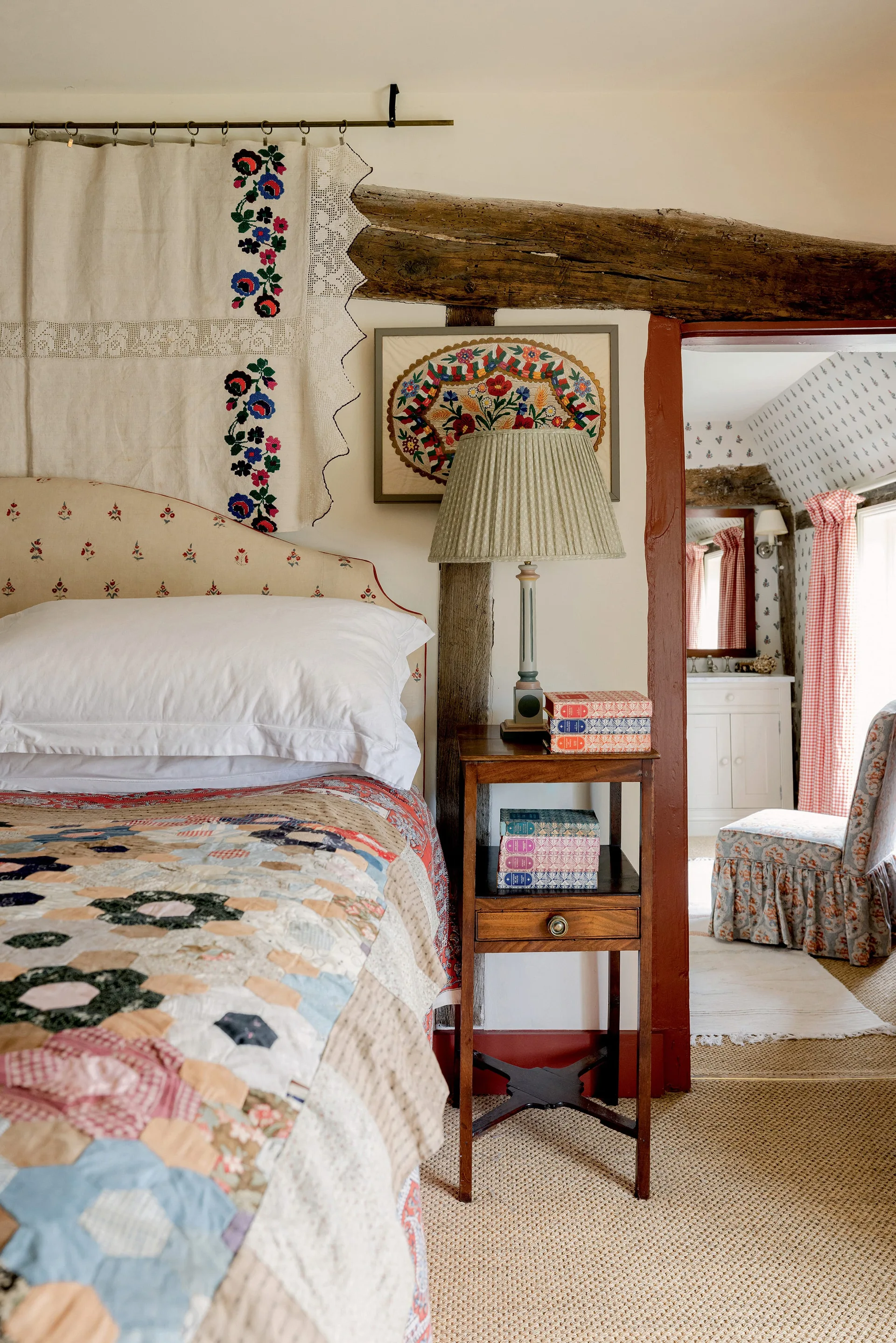 A cottage bedroom features a bed with a patchwork quilt, an upholstered headboard, an exposed ceiling beam, and a wooden nightstand next to a doorway.