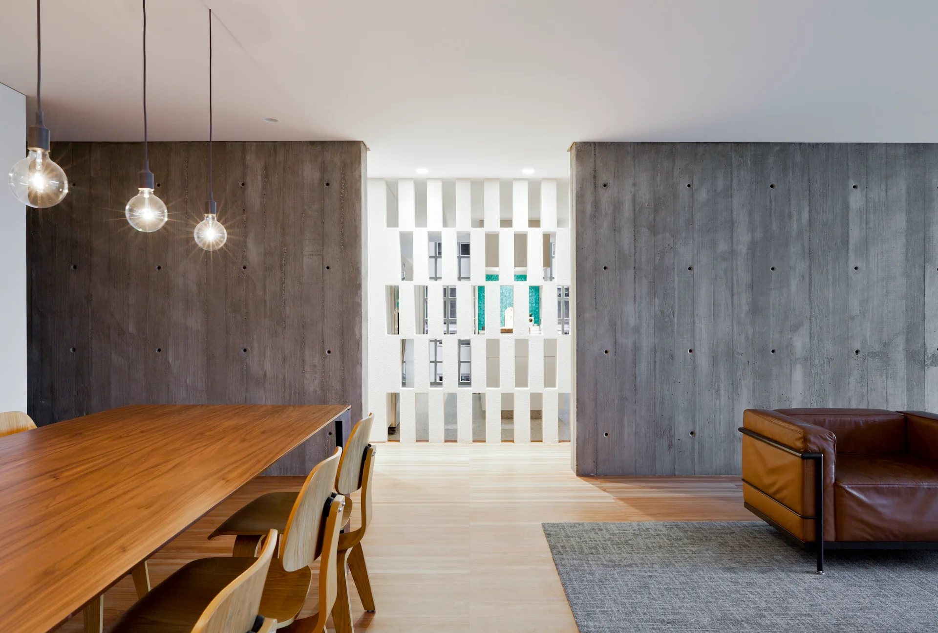 An open dining area features a wooden table, board-formed concrete walls, glass globe pendant lights, and a white geometric room divider.