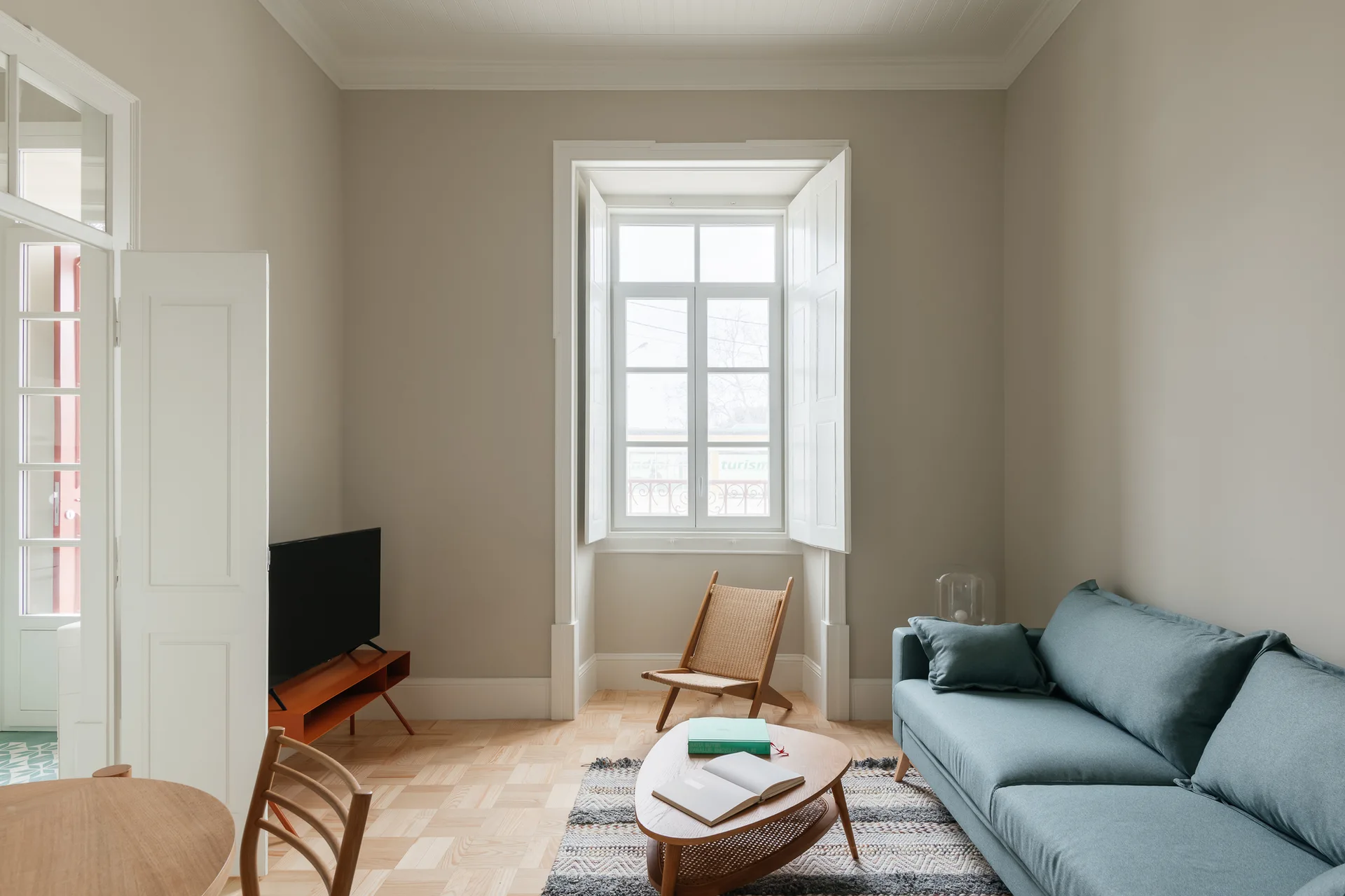 A light-filled beige living room featuring a blue sofa, a wooden coffee table, and a tall window with white folding shutters over parquet floors.