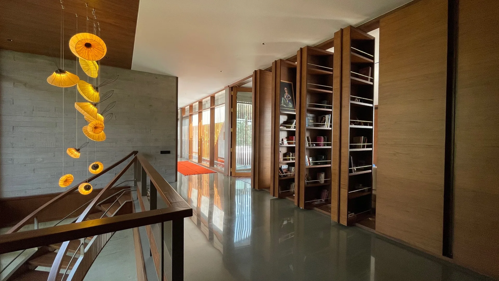 A modern hallway features a series of tall wooden pivoting bookshelves on one side, a reflective grey floor, and cascading orange pendant lights.