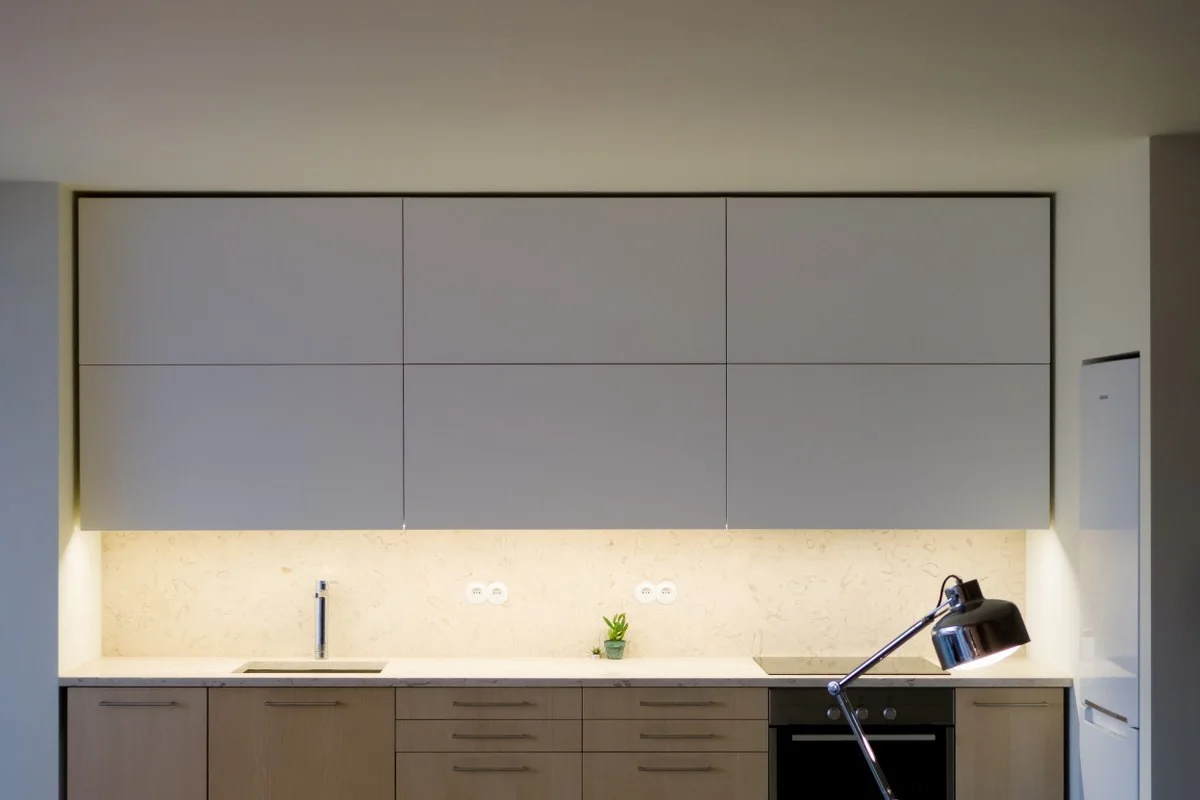 Minimalist kitchen featuring flat-front white upper cabinets, light wood base cabinets, and a stone backsplash illuminated by warm under-cabinet lighting.