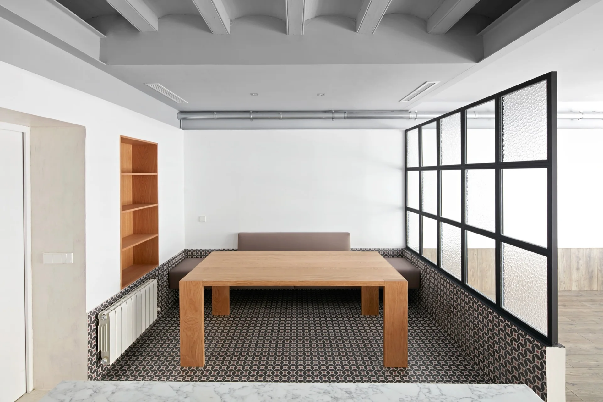 A modern dining nook features a solid wood table surrounded by built-in bench seating, geometric floor tile, and a black steel-framed glass partition.