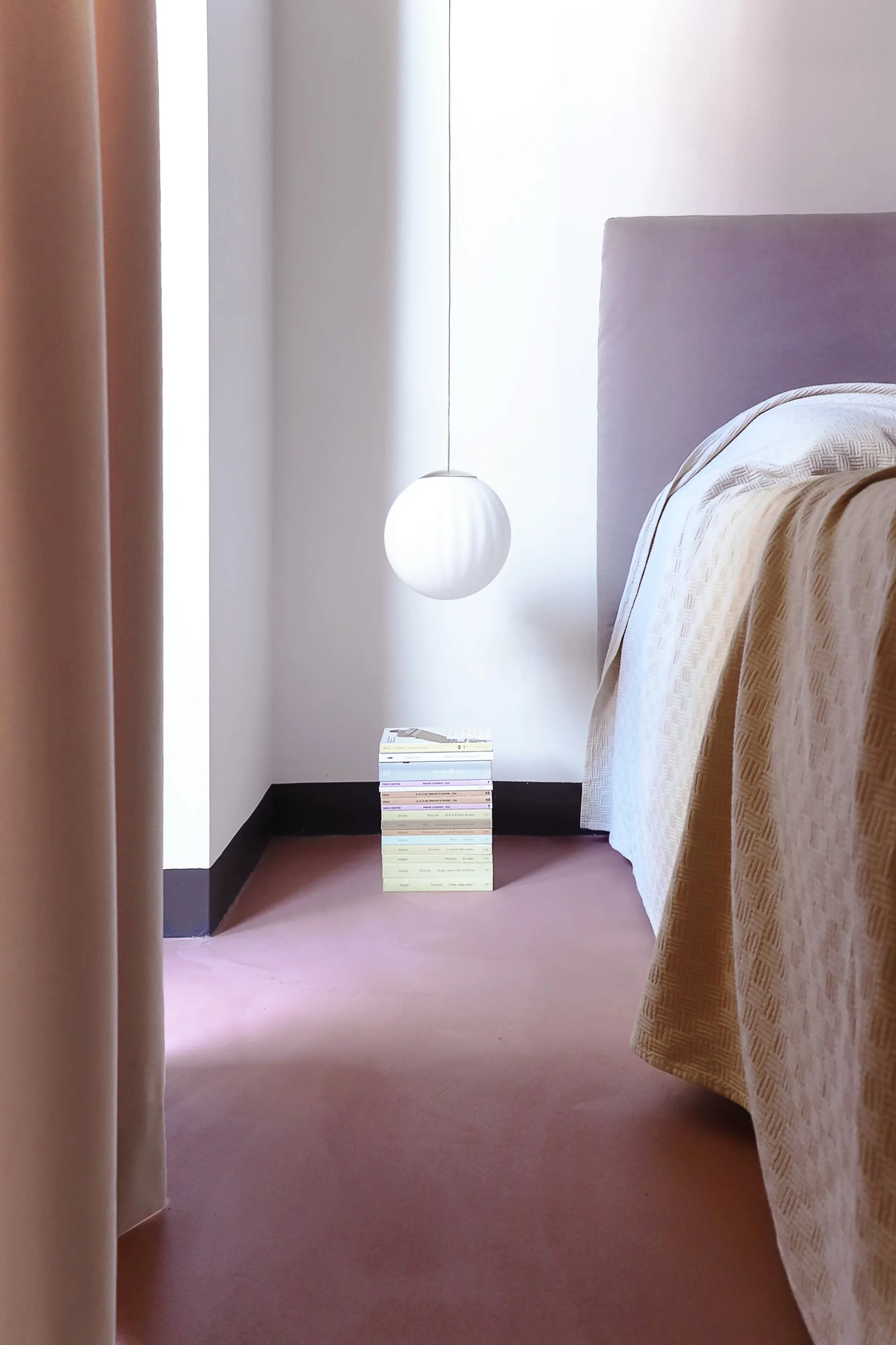 A minimalist bedroom corner featuring a spherical pendant light hanging above a neat stack of books next to an upholstered bed.
