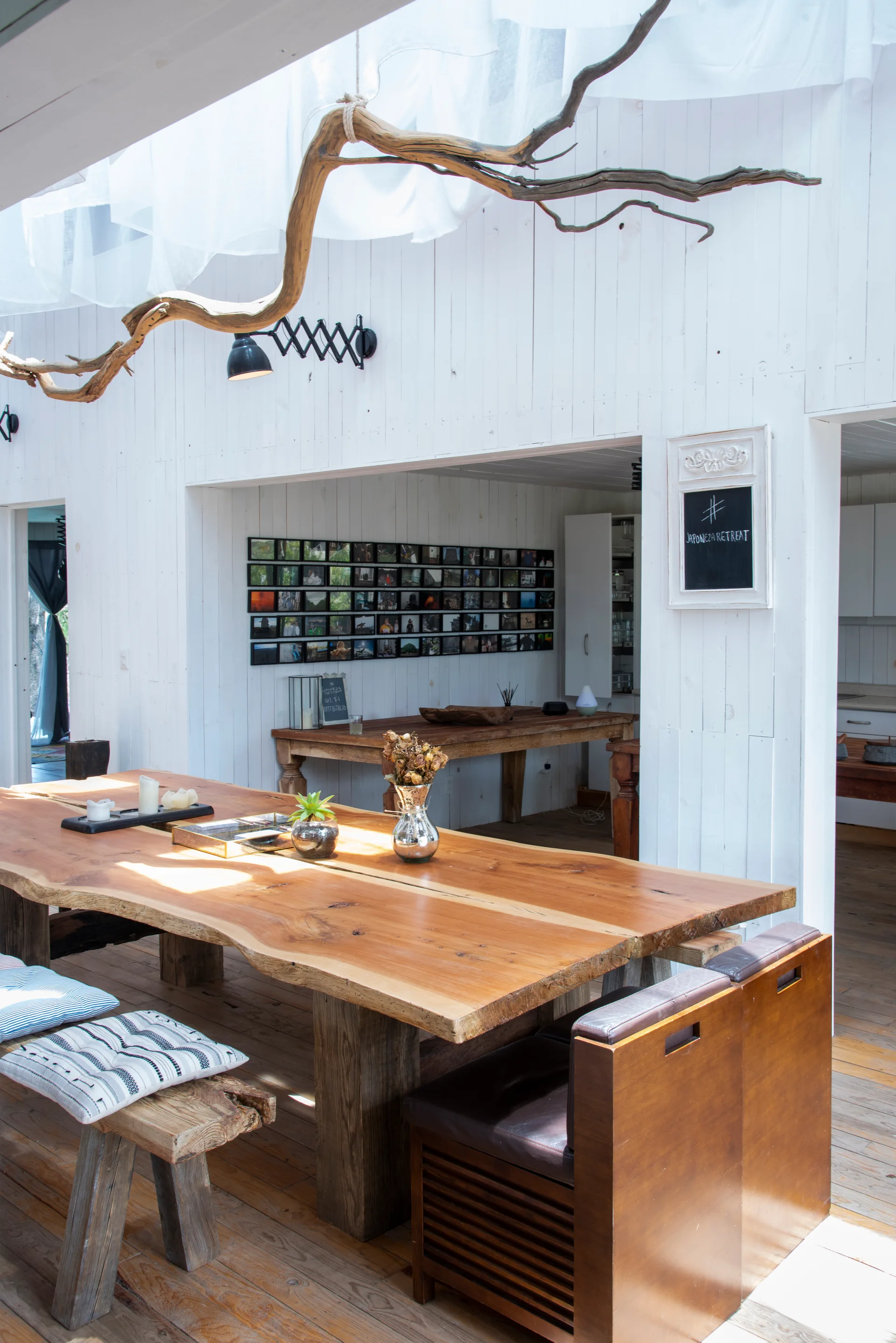 A rustic dining area features a long live edge wood dining table with benches, white paneled walls, and a large decorative branch hanging from the ceiling.