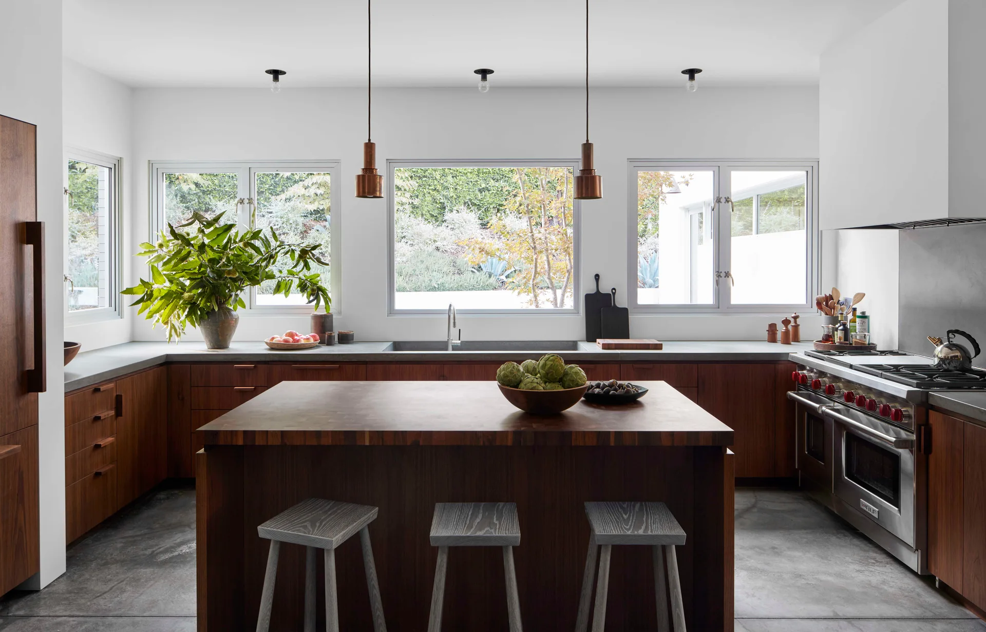A modern kitchen with dark wood cabinets, a central island with grey stools, concrete countertops, and three large windows overlooking a garden.