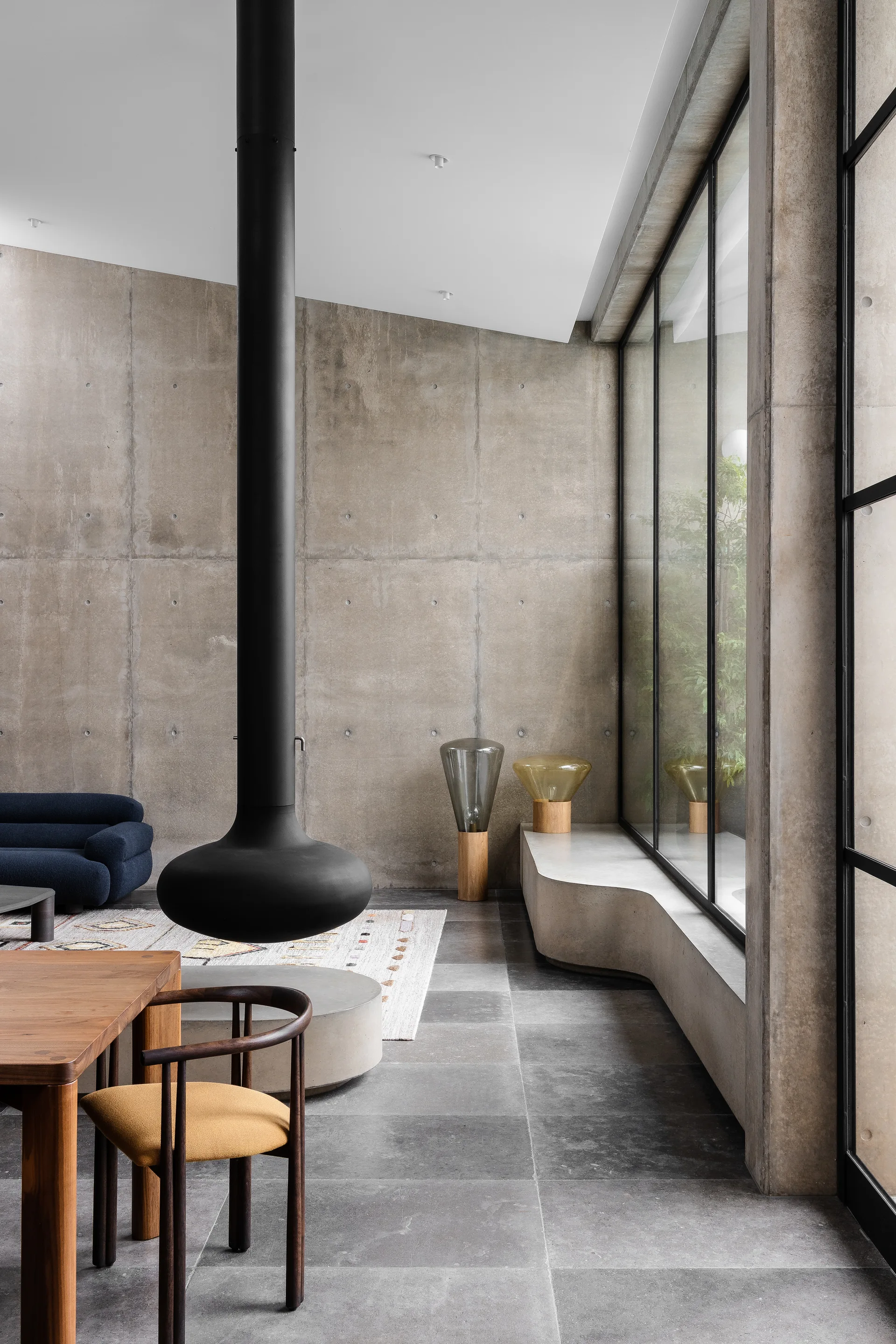 A minimalist room featuring a black suspended fireplace, exposed concrete walls, a curved concrete bench, and large metal-framed windows.